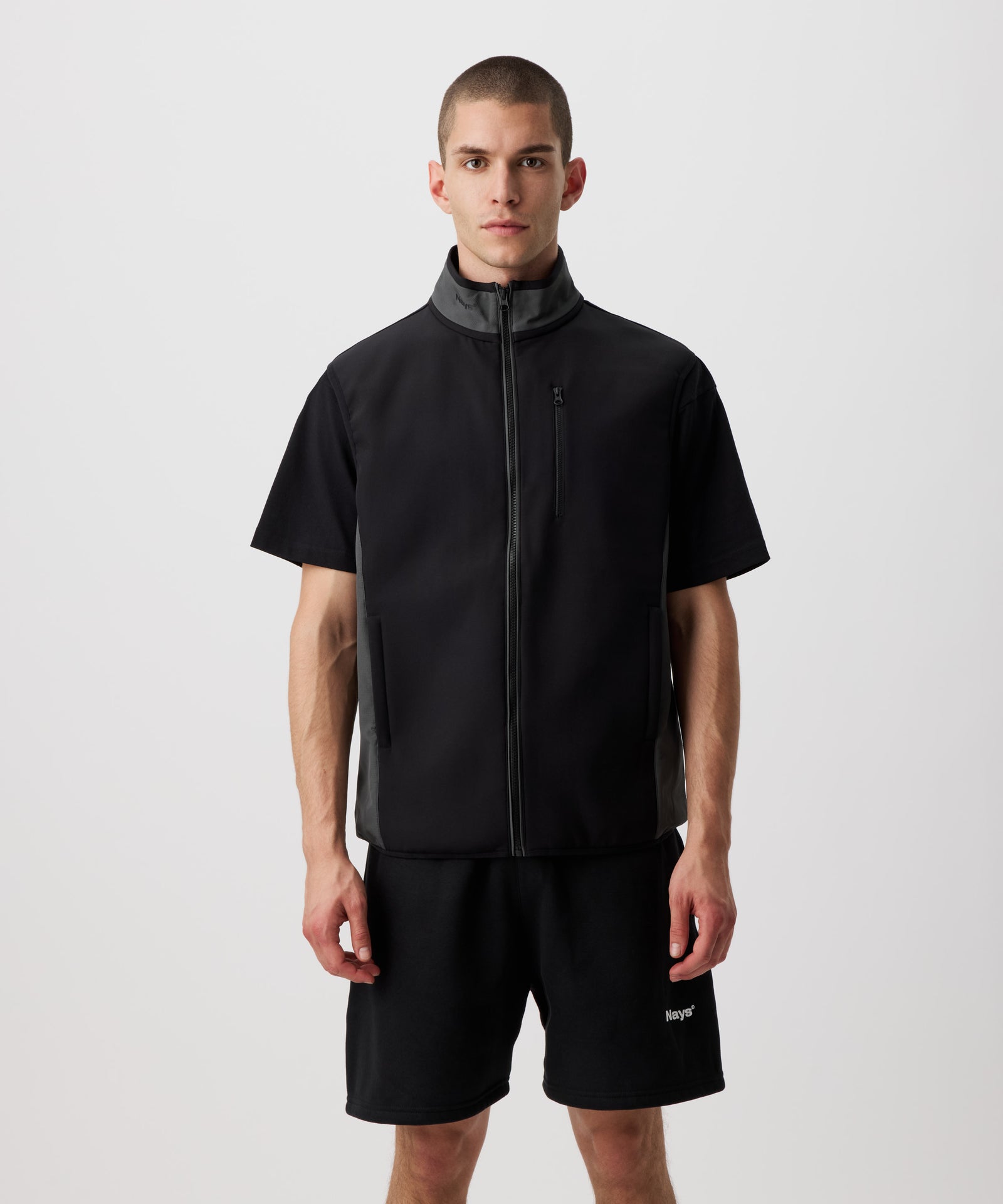 Ein Mann trägt eine dünne, funktionelle Weste in schwarz. Der Reißverschluss, die Seiten und der Kragen sind farblich in grau abgesetzt. Dazu trägt er eine schwarze Shorts.