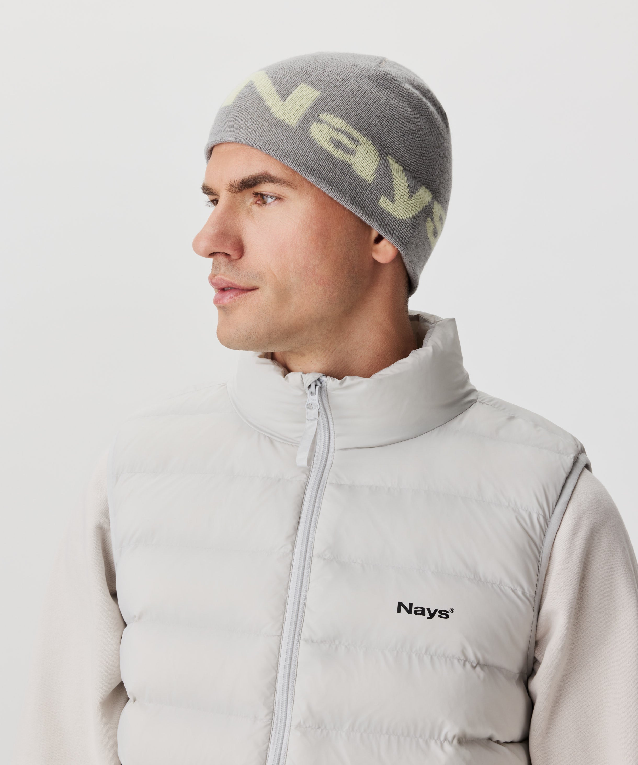 Ansicht im Halbseitenprofil Mann mit grauer Strickmütze und farblich abgesetztem Nays-Logo. Er trägt einen beige-farbigen Pullover und eine hell-graue Weste mit schwarzem Logo auf der linken Brust.
