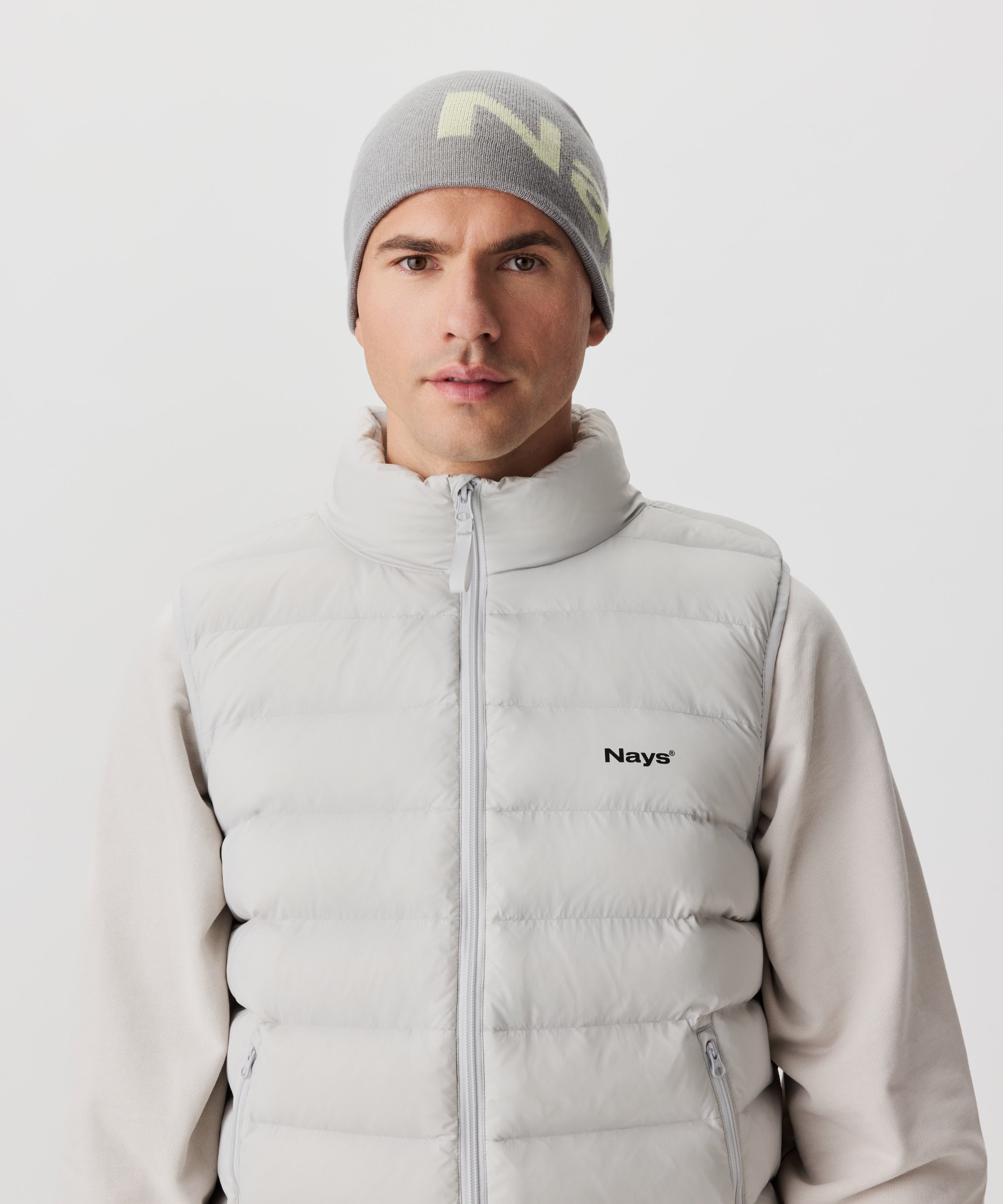 Frontansicht Mann mit grauer Strickmütze und farblich abgesetztem Nays-Logo. Er trägt einen beige-farbigen Pullover und eine hell-graue Weste mit schwarzem Logo auf der linken Brust.