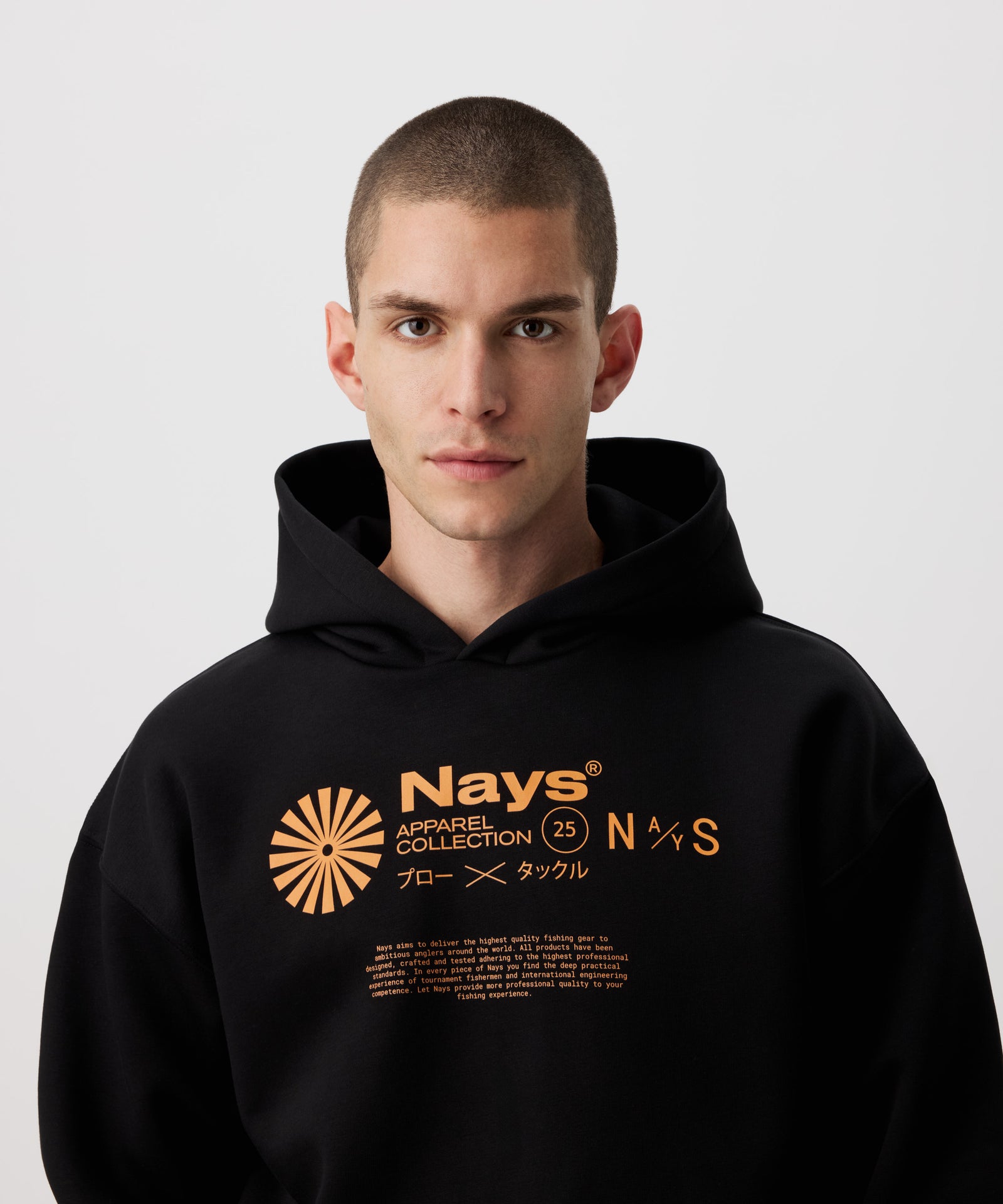 Eine Nahaufnahme des schwarzen Kapuzen-Pullover mit kontrastreichen Prints in einem leuchtenden Orange, getragen von einem Mann. Der Print dominiert durch ein Nays Schriftzug, mit weiteren Colelctionsangaben wie: Apparel Collection und "25", sowie ein Sonnen-ähnliches Symol.