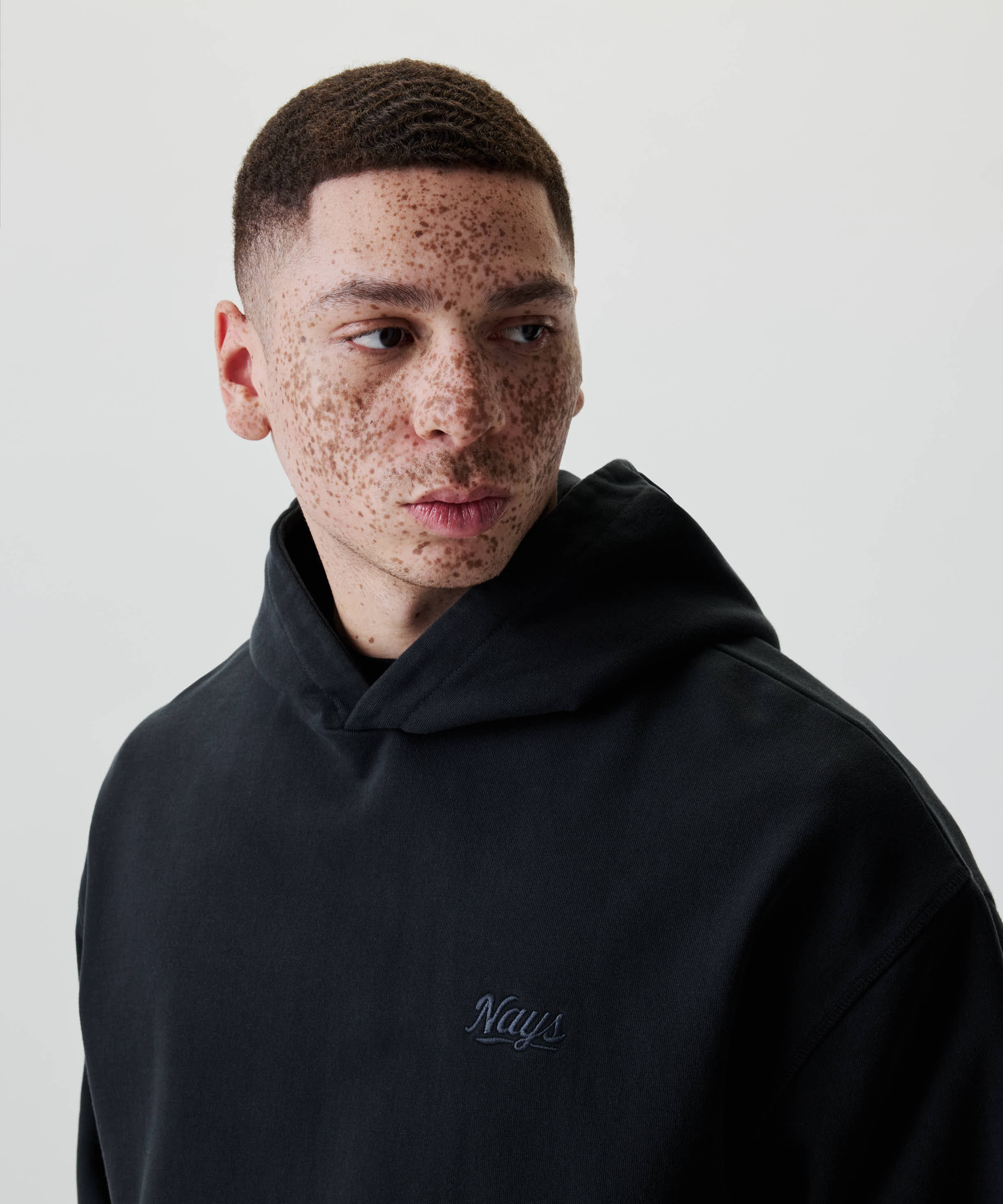 Junger Mann im Nays New Script Hoodie mit gesticktem Logo, blickt zur Seite. Der Hoodie bietet lässigen Stil und Komfort aus 100% Baumwolle.