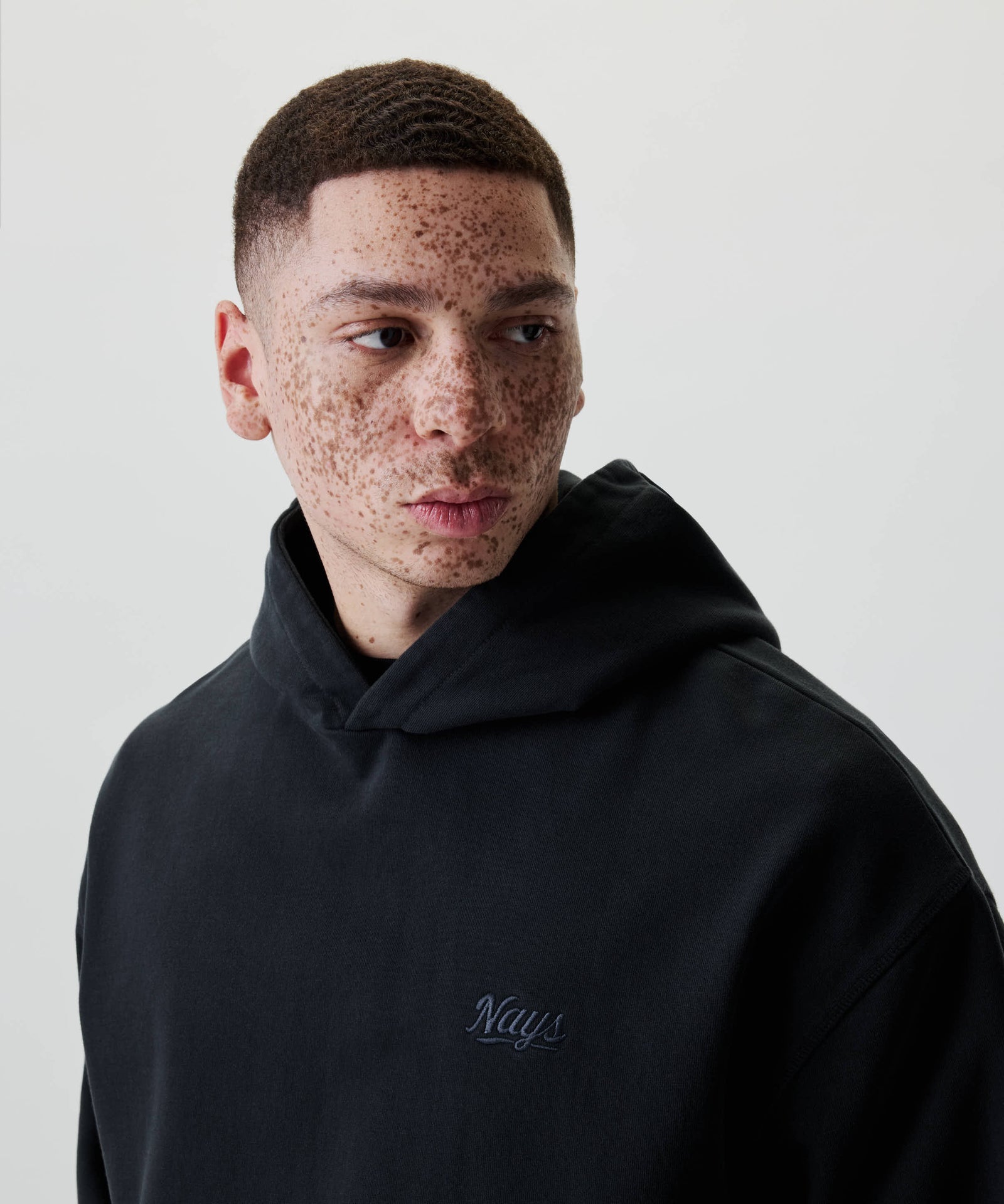 Junger Mann im Nays New Script Hoodie mit gesticktem Logo, blickt zur Seite. Der Hoodie bietet lässigen Stil und Komfort aus 100% Baumwolle.