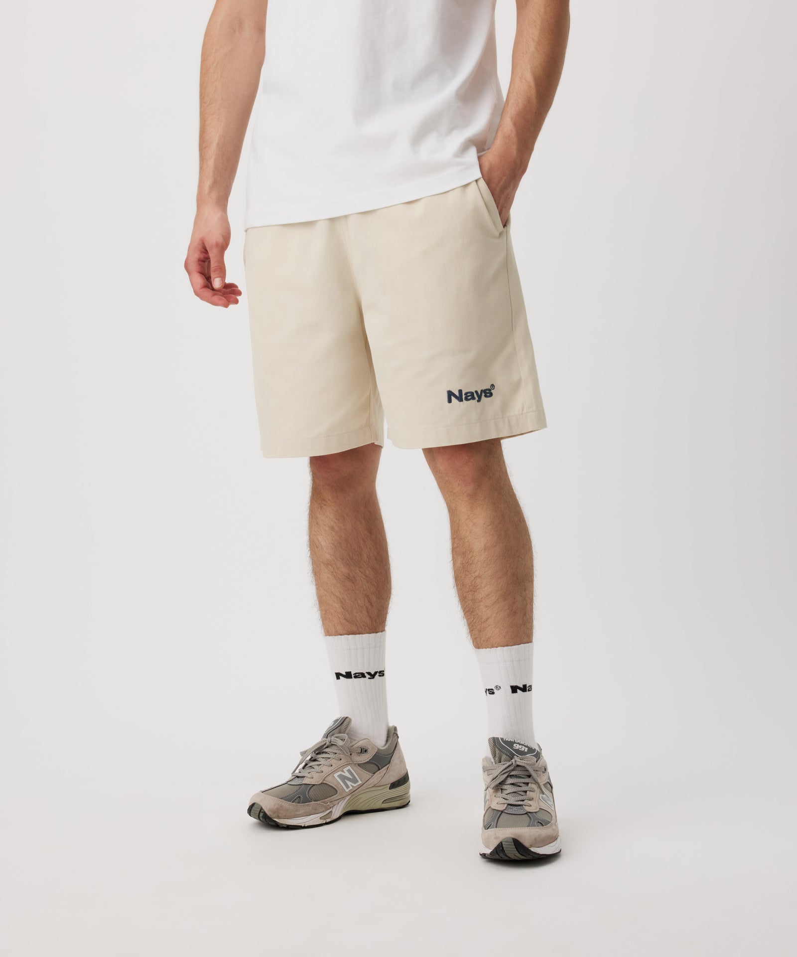 Ein Mann steht schräg zur Kamera. Er trägt ein weißes T-Shirt, eine beige Shorts mit einem kleinen, grünen Logo auf dem linken Bein. Dazu trägt er weiße Logo Socken und Sportschuhe.