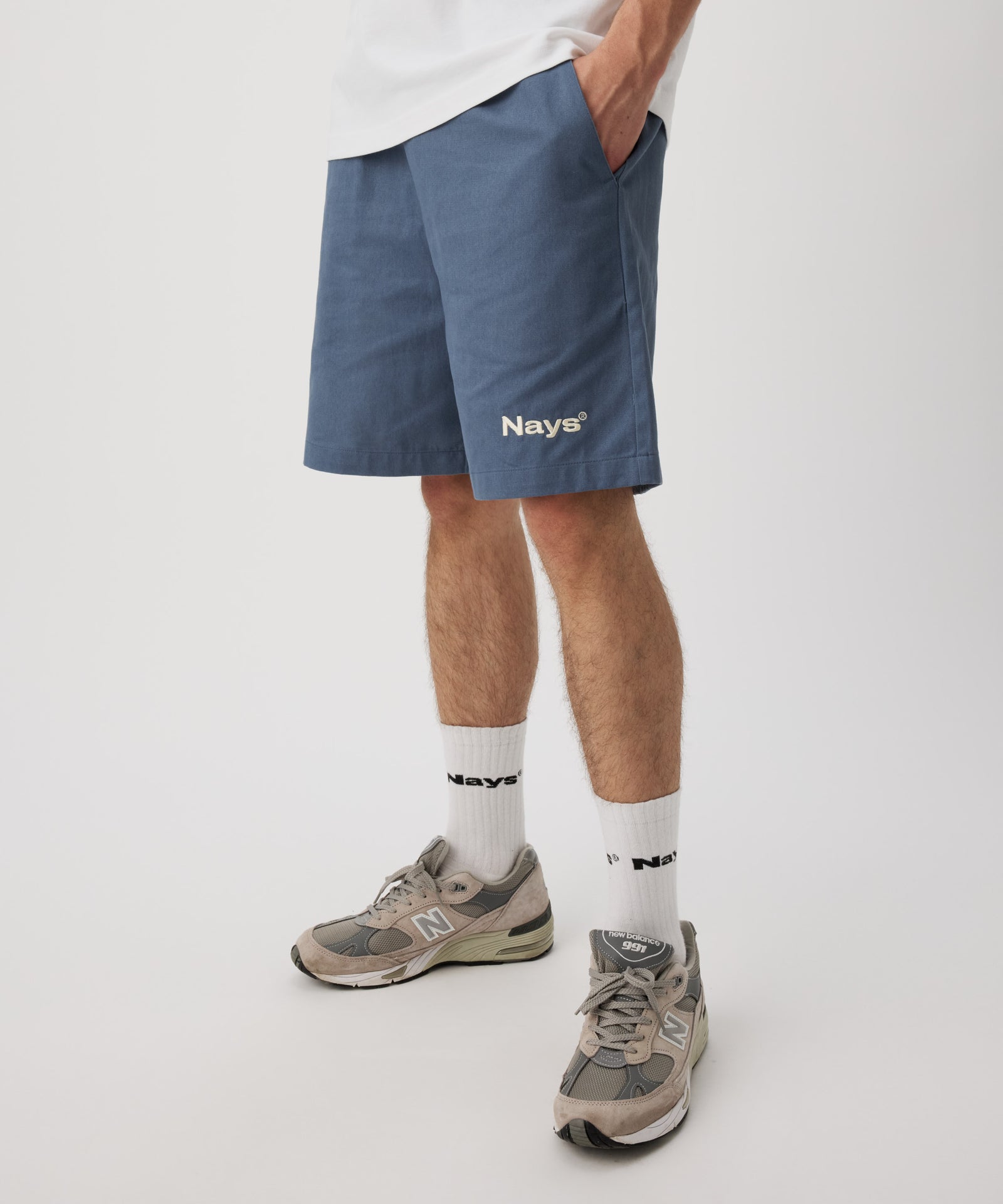 Ein Mann trägt eine blaue Shorts mit einem weißen "Nays"-Logo auf dem linken Bein und hat seine linke Hand in der Tasche. Dazu trägt er ein weißes T-Shirt und eine weiße Socken.