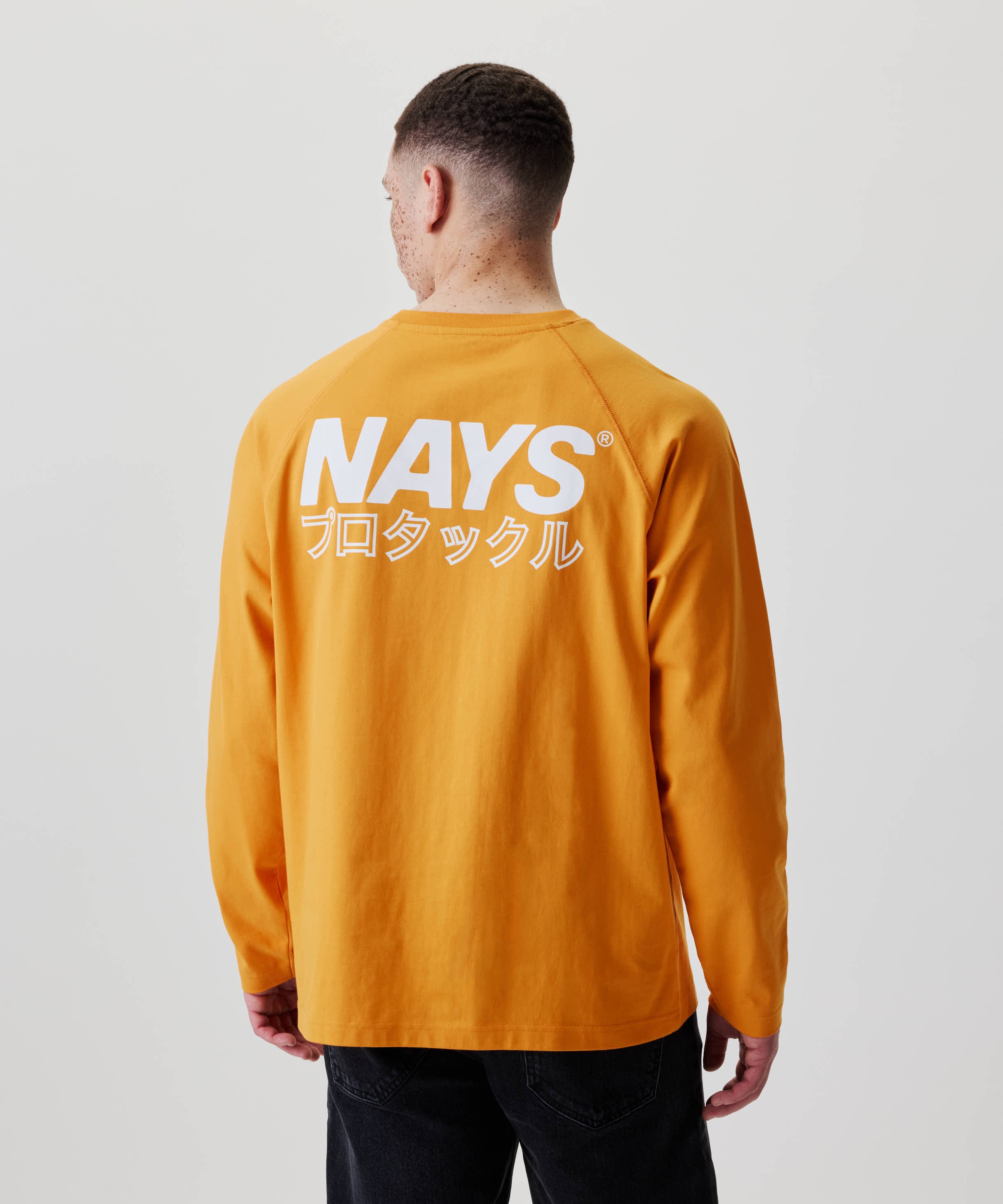 Mann von hinten im Nays Pro Tackle International Longsleeve mit NAYS und japanischem Text auf dem Rücken, kombiniert mit dunklen Jeans.