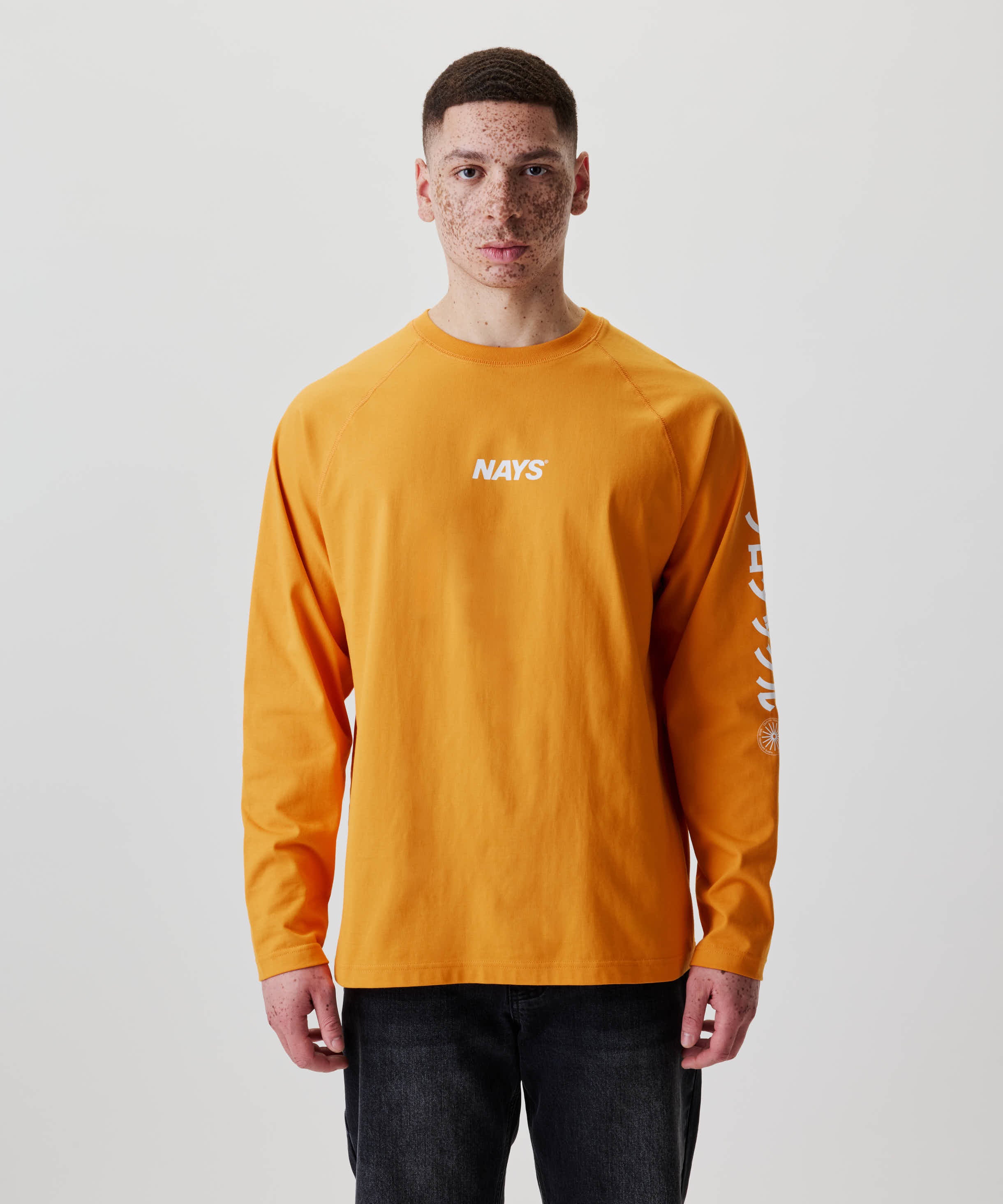 Person trägt das Nays Pro Tackle International Longsleeve mit weißem NAYS-Aufdruck auf der Brust und zusätzlichem Print auf einem Ärmel, kombiniert mit dunklen Hosen.