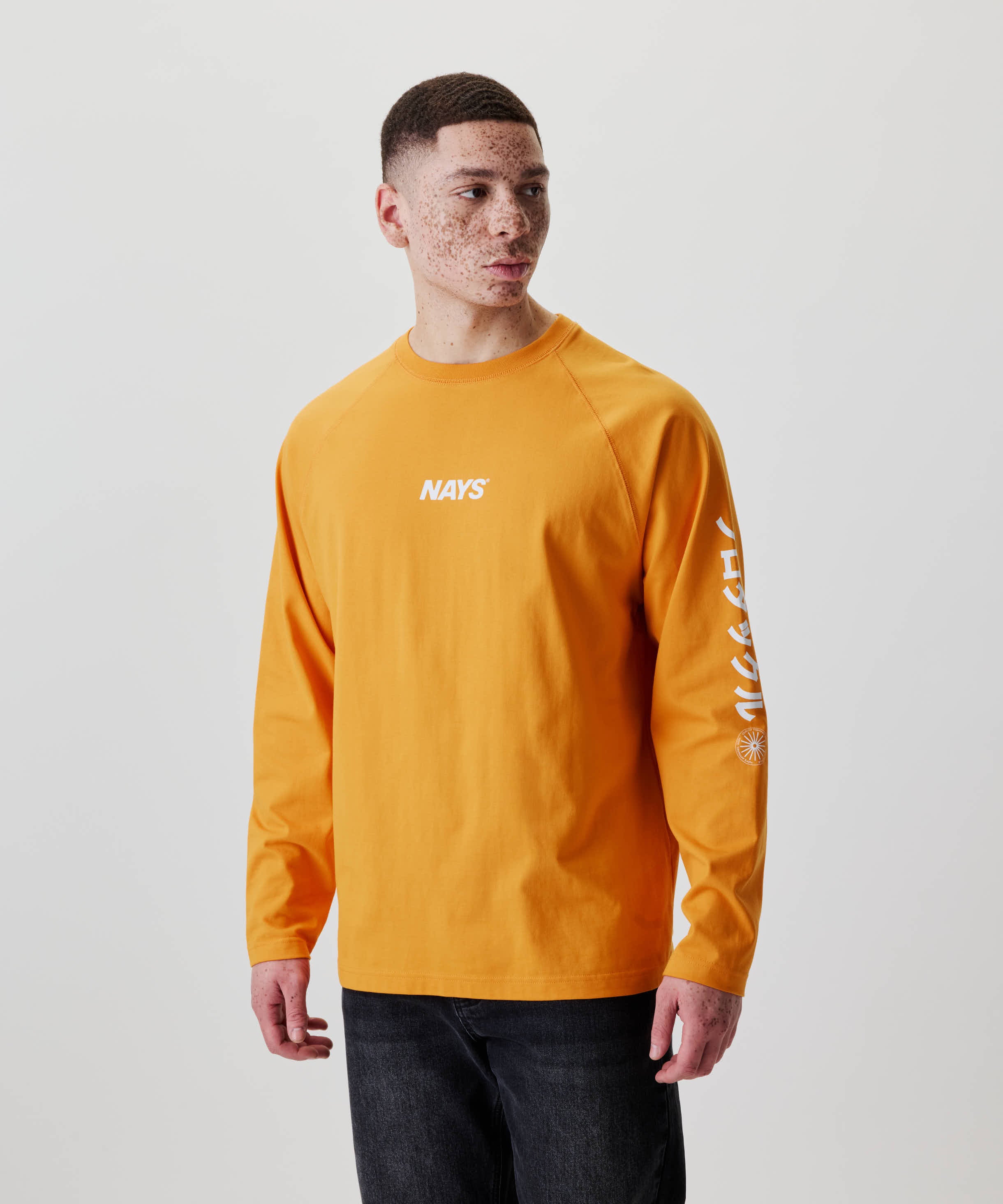 Eine junge Person trägt das Nays Pro Tackle International Longsleeve mit NAYS-Aufdruck auf der Brust und grafischen Buchstaben am Ärmel, kombiniert mit dunkler Hose.