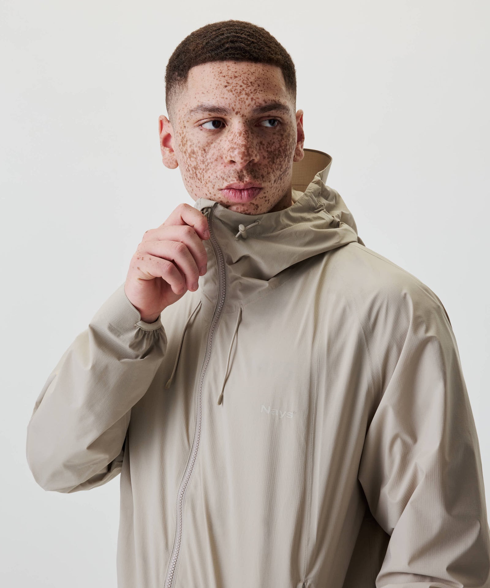 Junger Mann trägt Nays 2L Defend Mono Jacket, zieht Kapuze hoch; leichte, wasserdichte Jacke mit Raglanärmeln und elastischen Bündchen für Bewegungsfreiheit.
