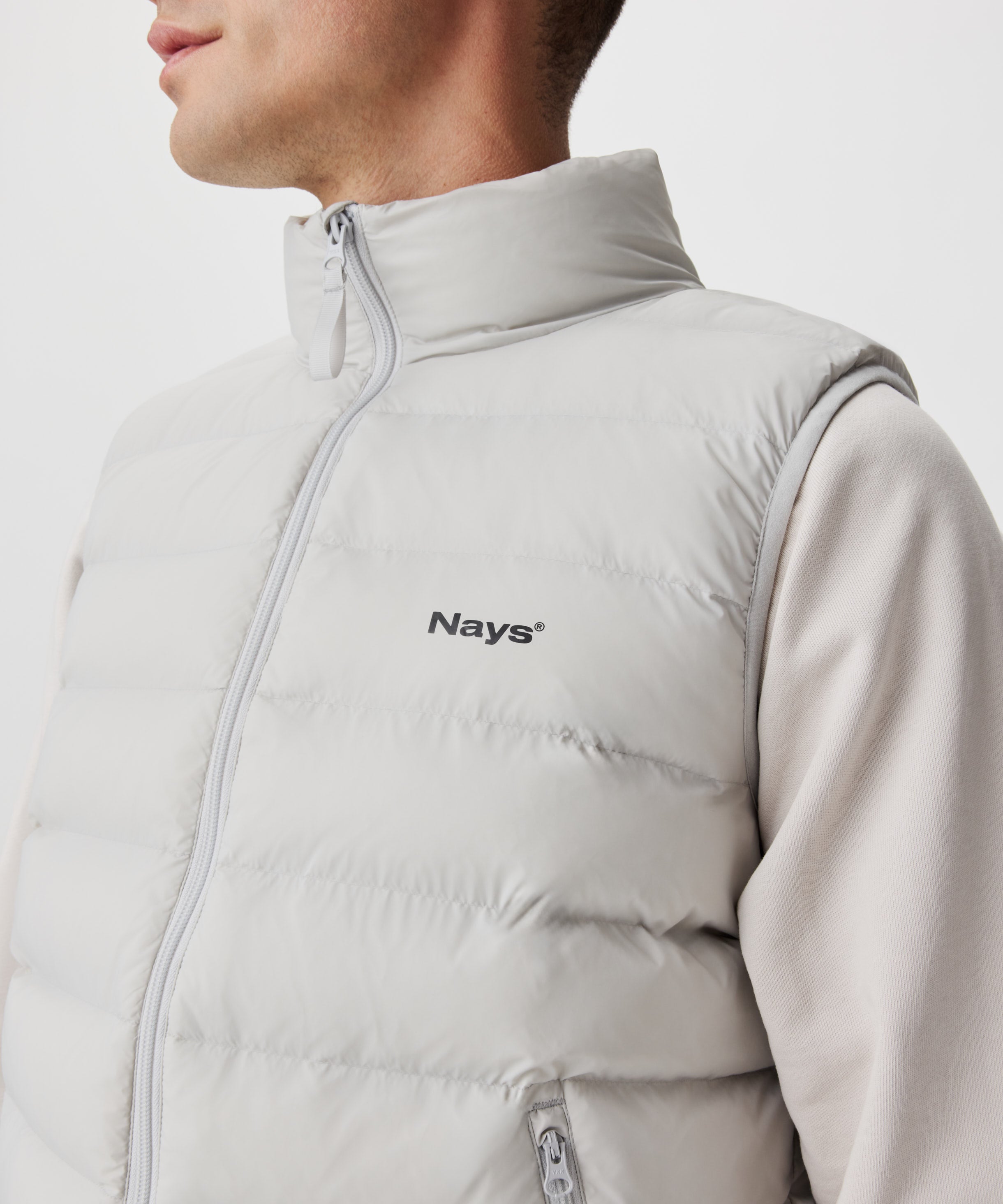 Mann in hell-grauer Weste. Die Weste verfügt über einen tonalen Reißverschluss und ein dezentes, schwarzes Nays-Logo auf der linken Brust.