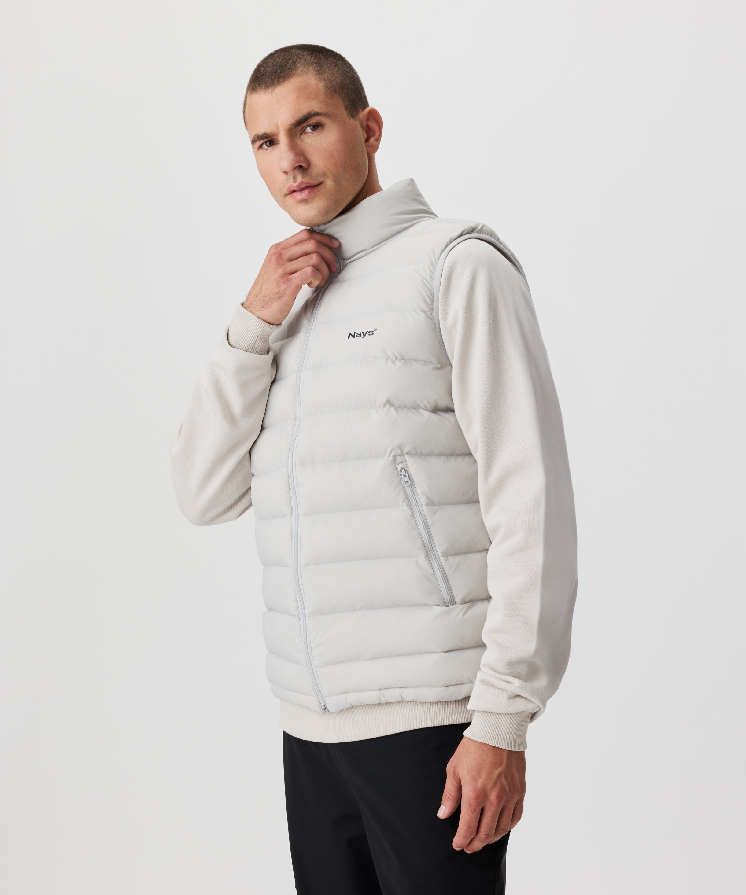 Mann in hell-grauer Weste und schwarzer Hose. Die Weste verfügt über einen tonalen Reißverschluss und ein dezentes, schwarzes Nays-Logo auf der linken Brust.