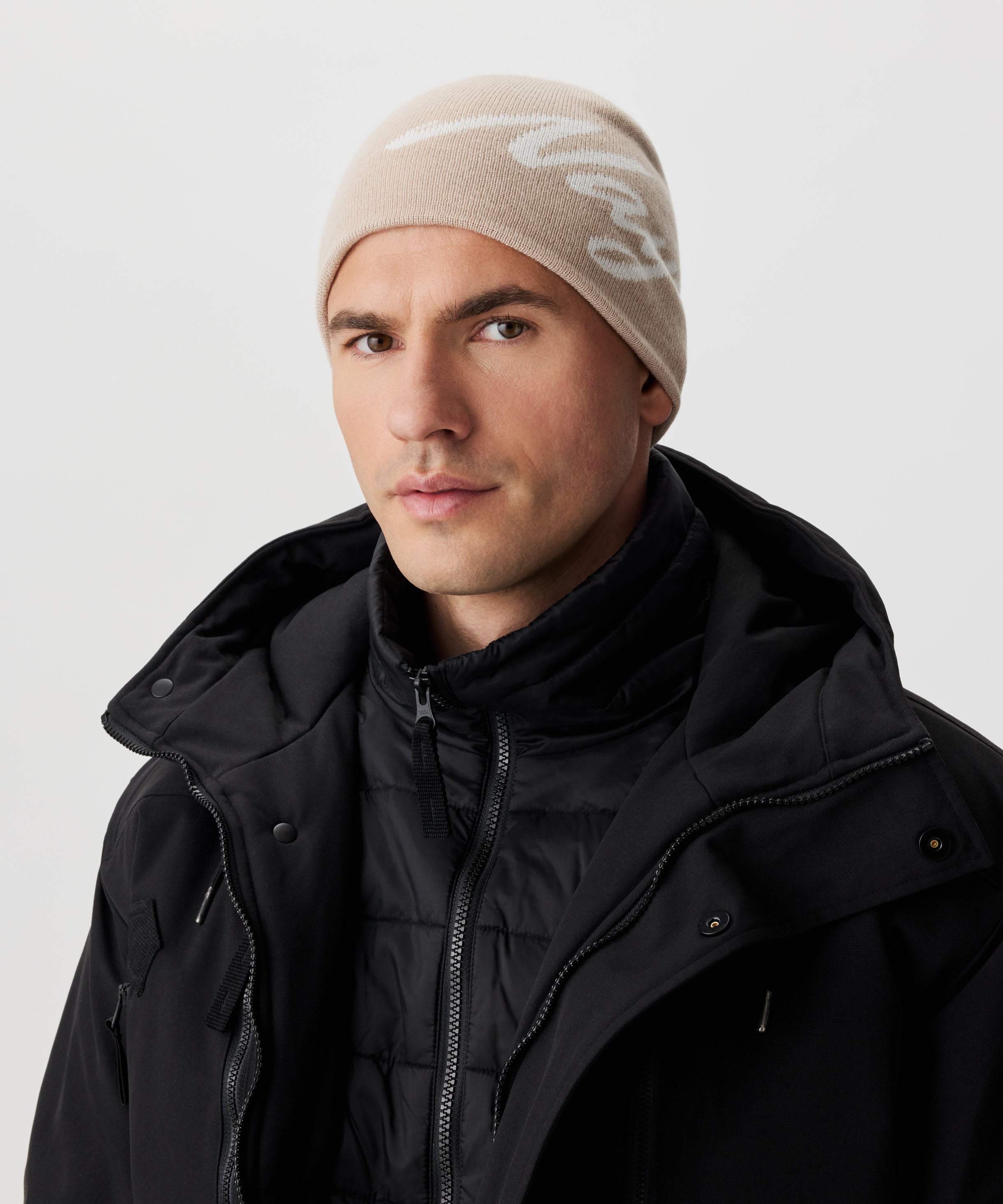 Frontale Ansicht Mann in schwarzer Jacke und beige-farbiger Strickmütze mit weißem Nays-Logo-Schriftzug.