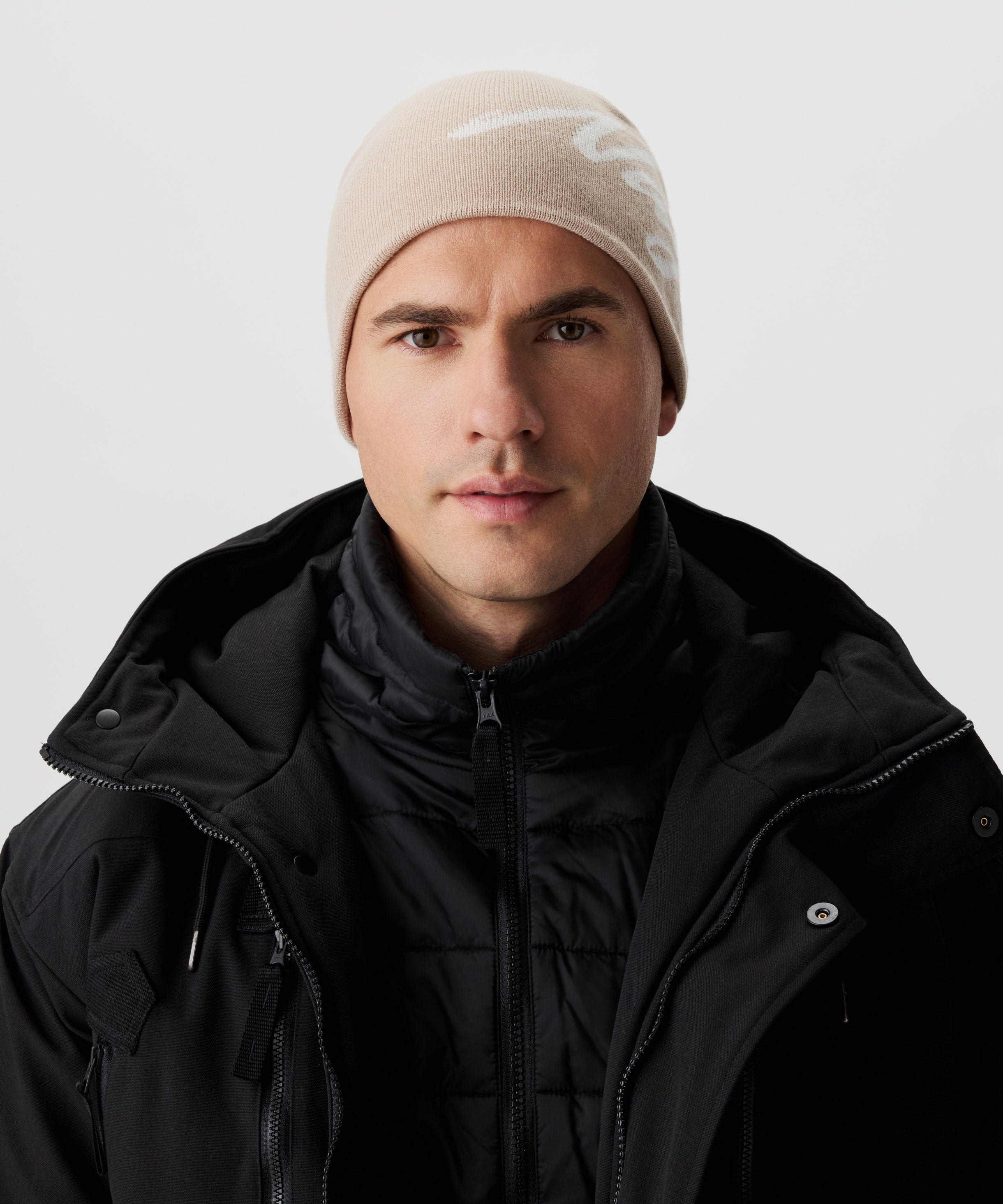 Frontale Ansicht Mann  in schwarzer Jacke und beige-farbiger Strickmütze mit weißem Nays-Logo-Schriftzug.