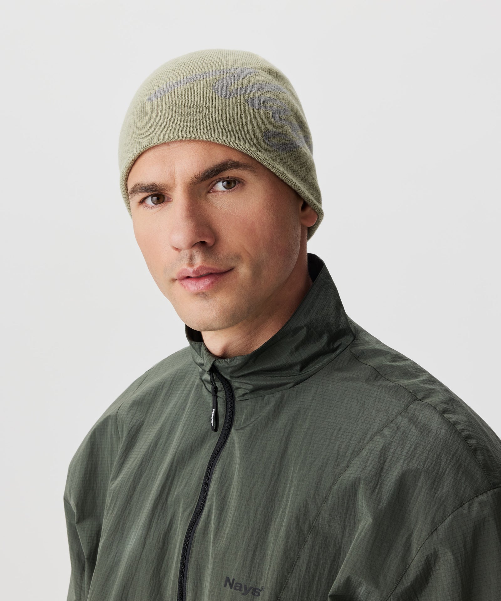 Frontale ansicht Mann in grüner Jacke und grüner Strickmütze mit grauen Nays-Logo-Schriftzug.