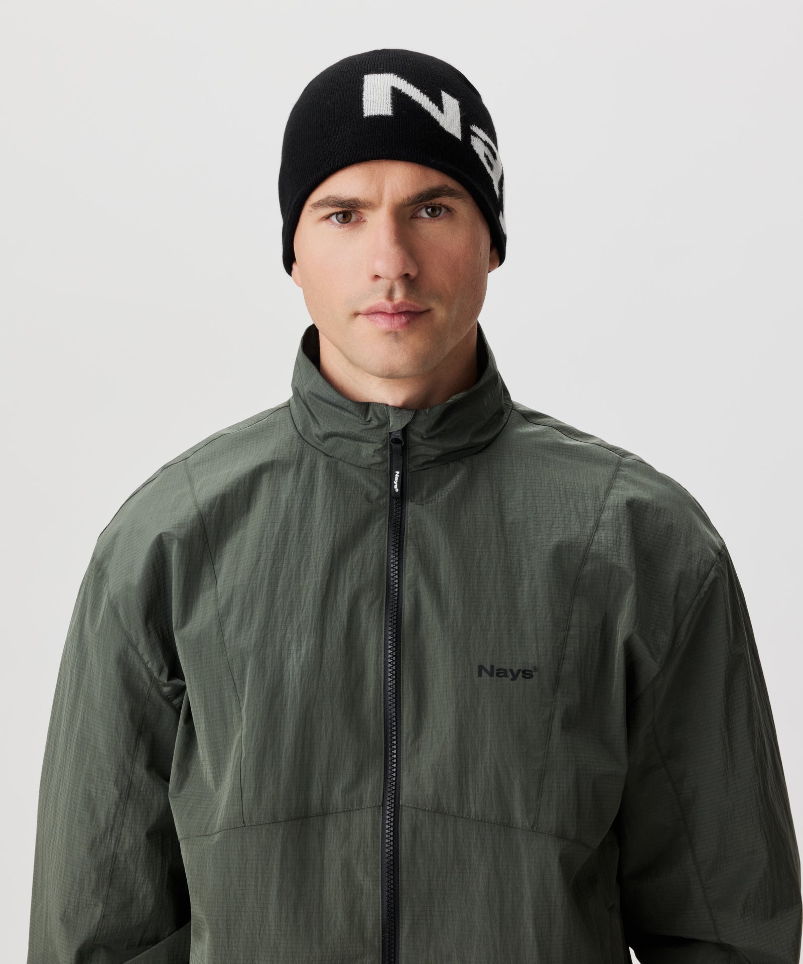 Frontale Ansicht Mann in grüner Jacke und schwarzer Strickmütze mit weißem Nays-Logo-Schriftzug.