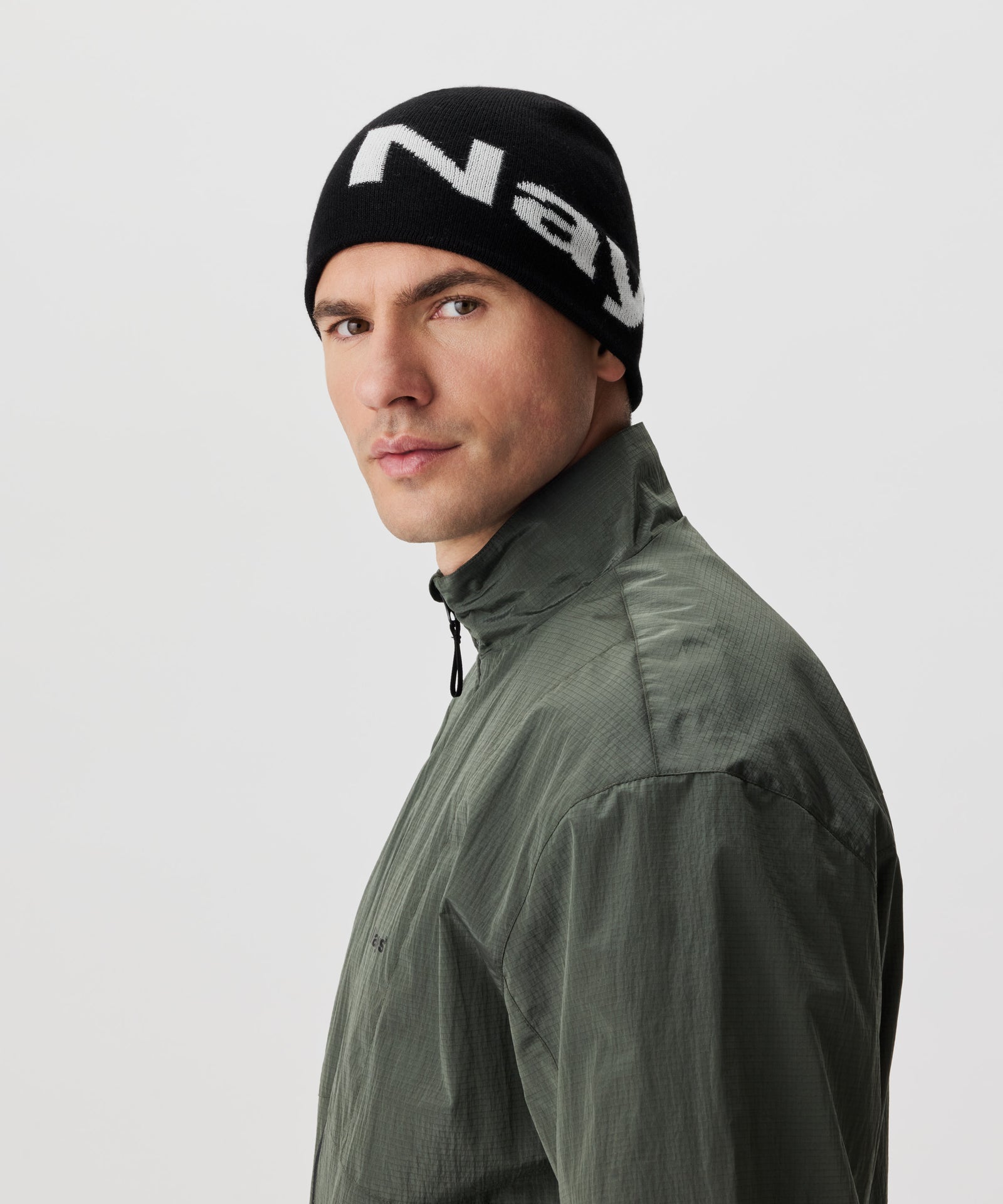 Halbseitliche Ansicht Mann in grüner Jacke und schwarzer Strickmütze mit weißem Nays-Logo-Schriftzug.