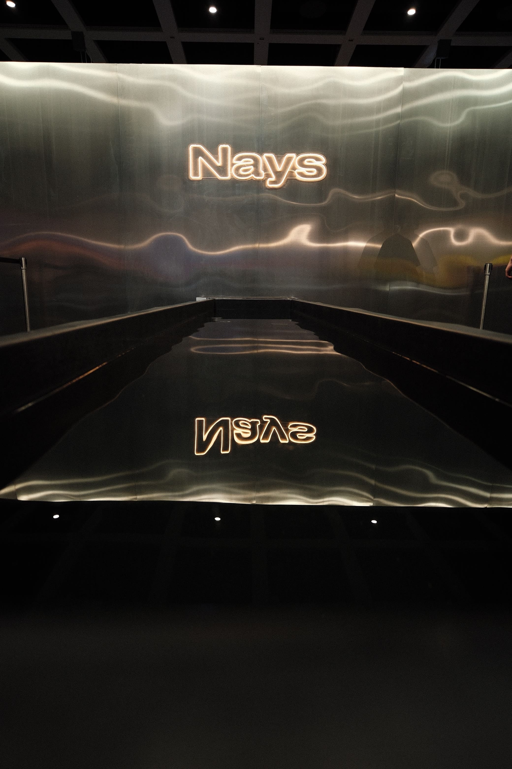 Beleuchteter Nays Markenschriftzug an einer metallischen Wand mit Spiegelung im Wasserbecken.
