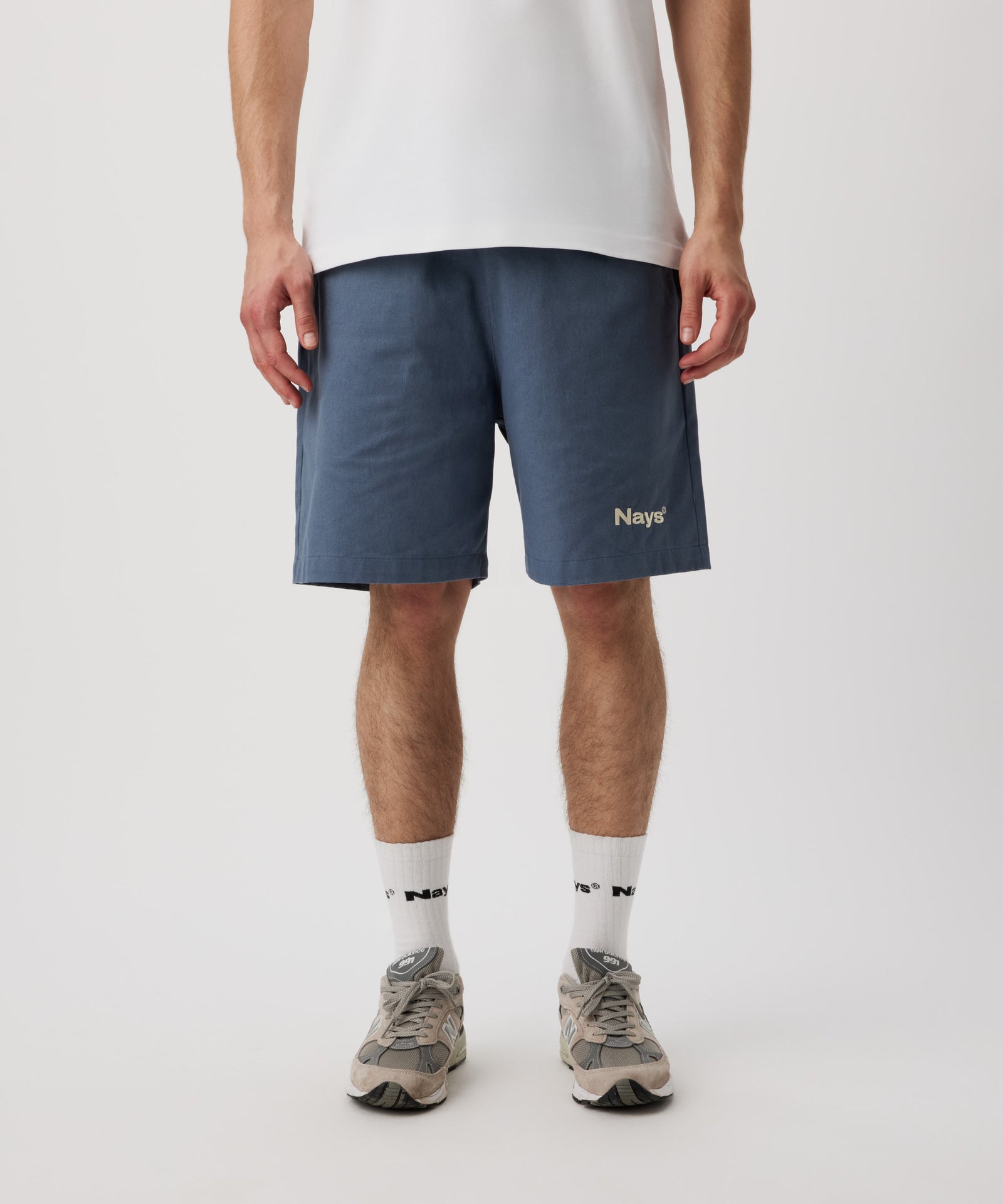Ein Mann trägt ein weißes T-Shirt, eine blaue Shorts mit einem kleinen, weißen Logo auf dem linken Bein. Dazu trägt er weiße Logo Socken und graue Sportschuhe.