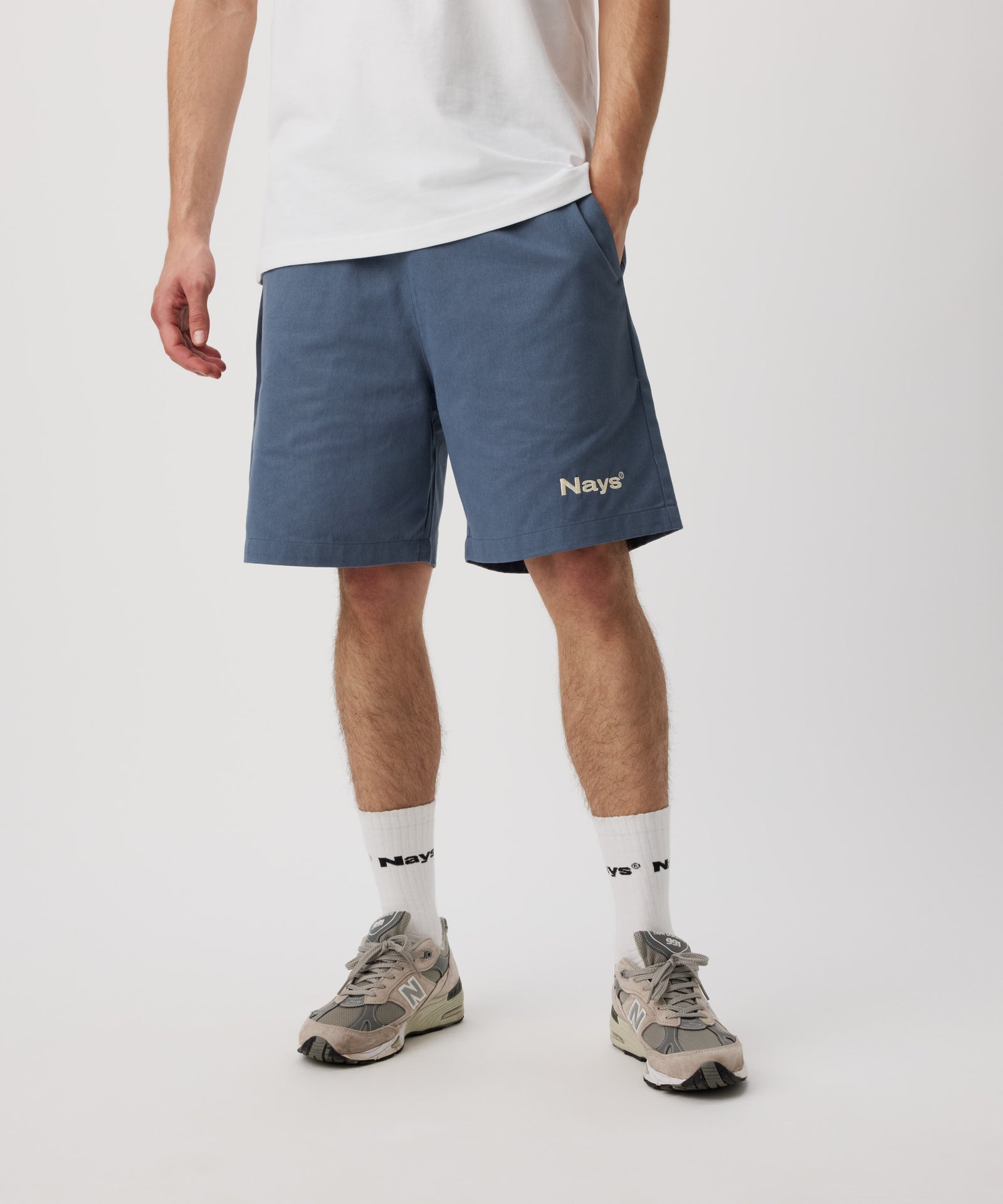 Ein Mann steht schräg zur Kamera. Er trägt ein weißes T-Shirt, eine blaue Shorts mit einem gestickten, weißen Logo auf dem linken Bein. Dazu trägt er weiße Logo Socken und beige Sportschuhe.