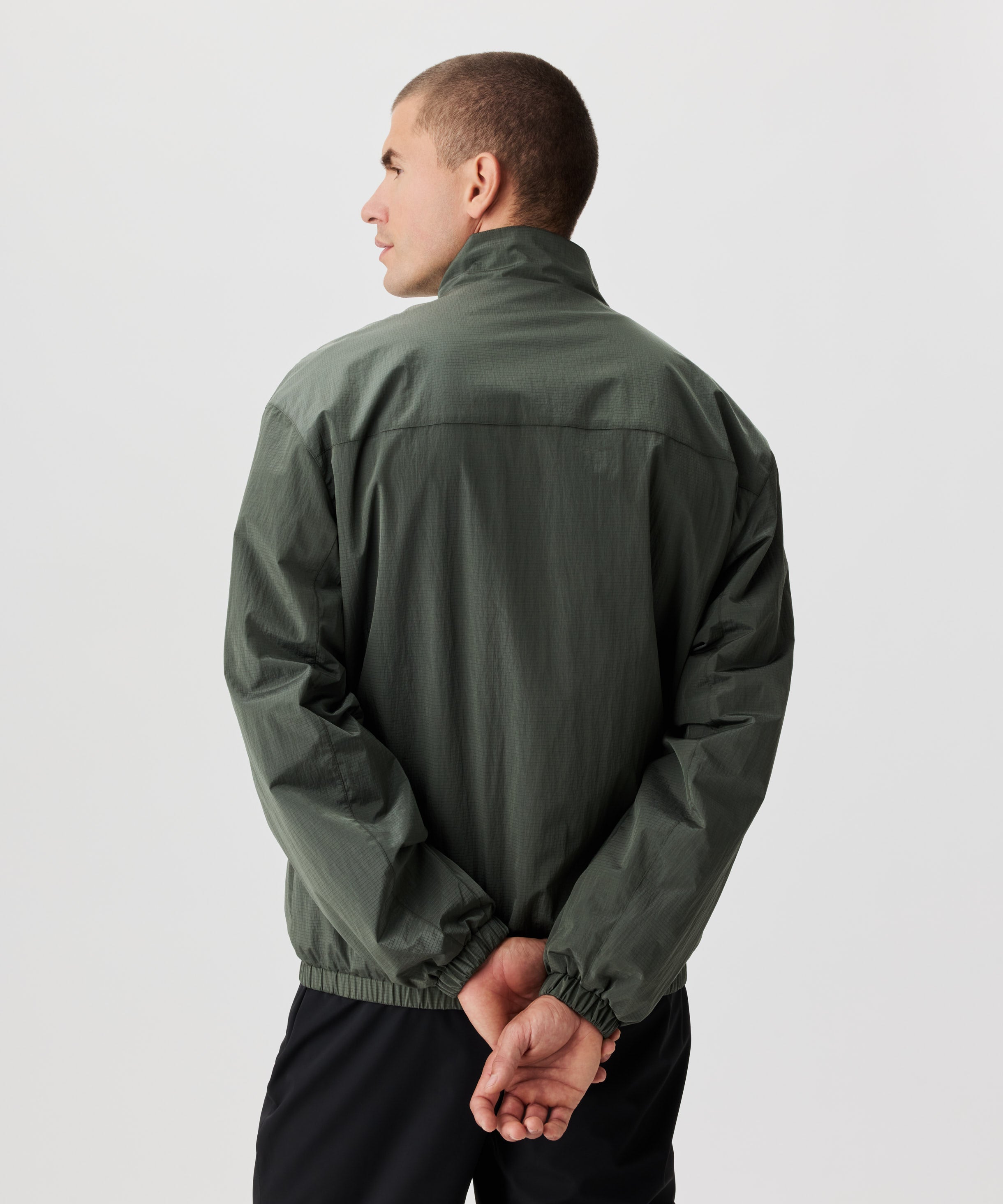 Mann in grüner Blouson Jacke in der Rückansicht. Die Arme sind hinter dem Rücken gekreuzt.