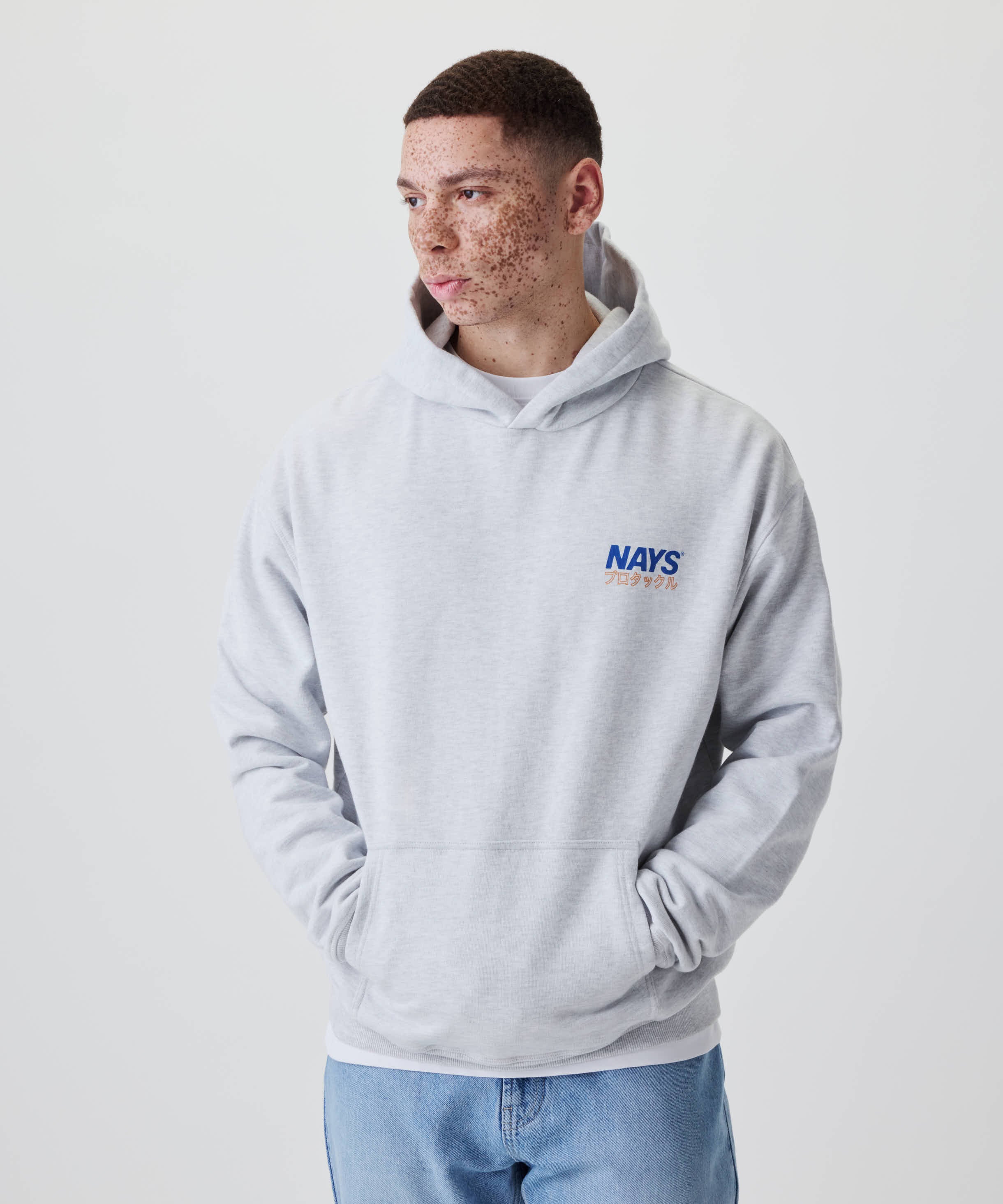 Junger Mann trägt Nays Pro Tackle International Hoodie mit kleinem Brustprint. Hände in der Kängurutasche, legerer Freizeitlook.