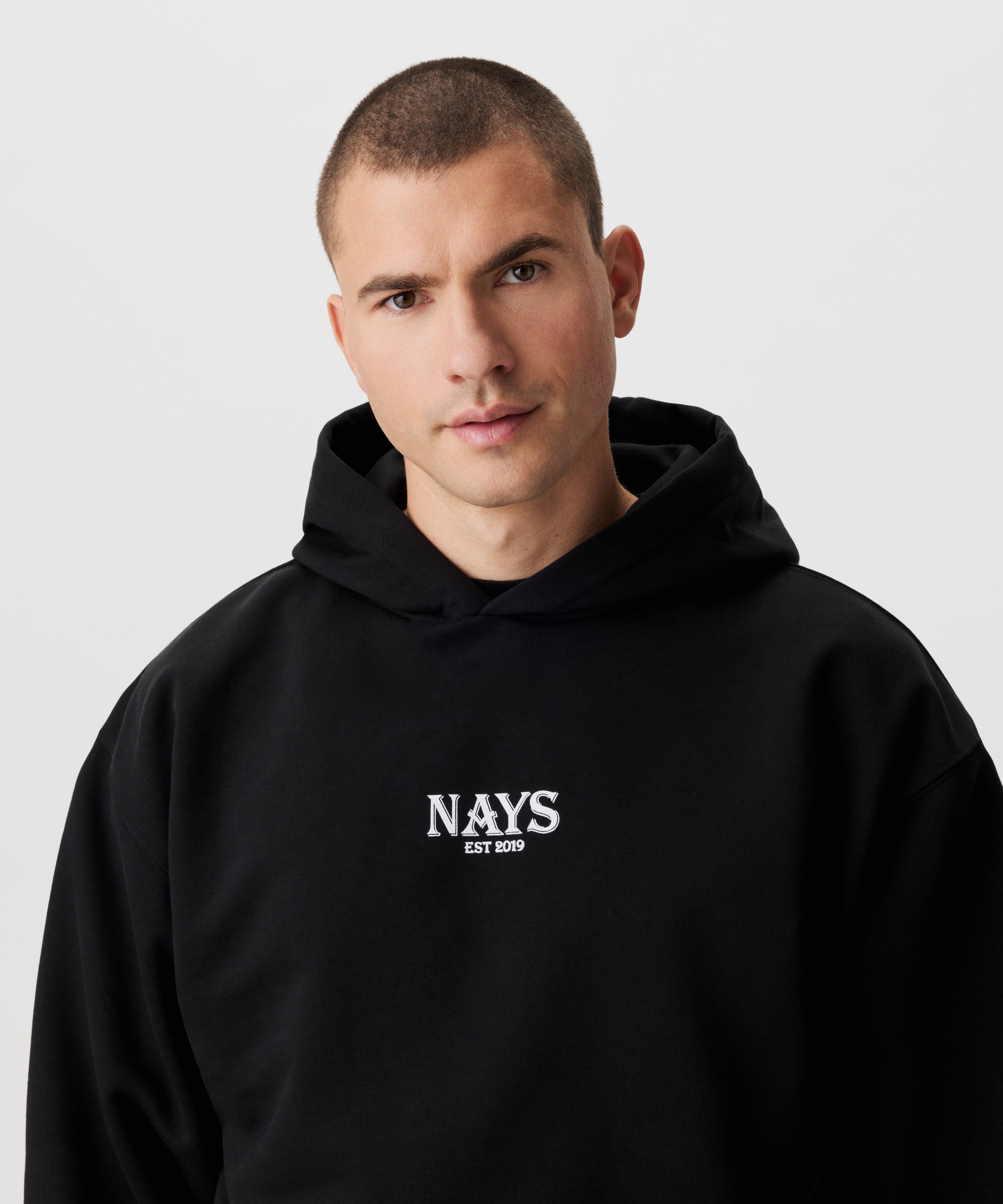 Frontansicht Mann im schwarzen Kapuzen Sweatshirt. Das Sweatshirt hat einen mittig gesetztes Nays-Logo in weiß.