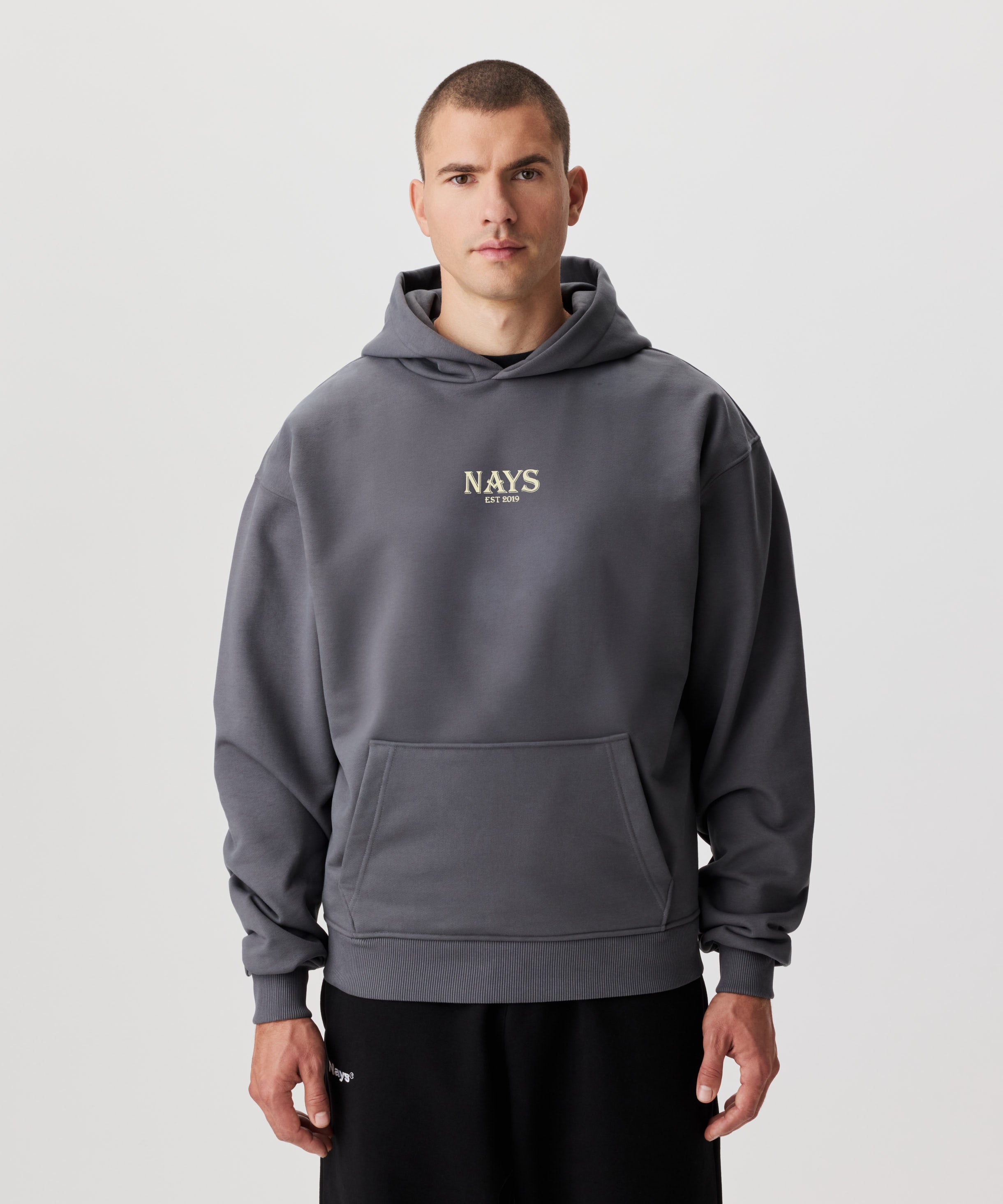 Mann im grauen Kapuzen Sweatshirt in der Frontansicht. Das Sweatshirt hat einen mittig gesetztes Nays-Logo in gelb.