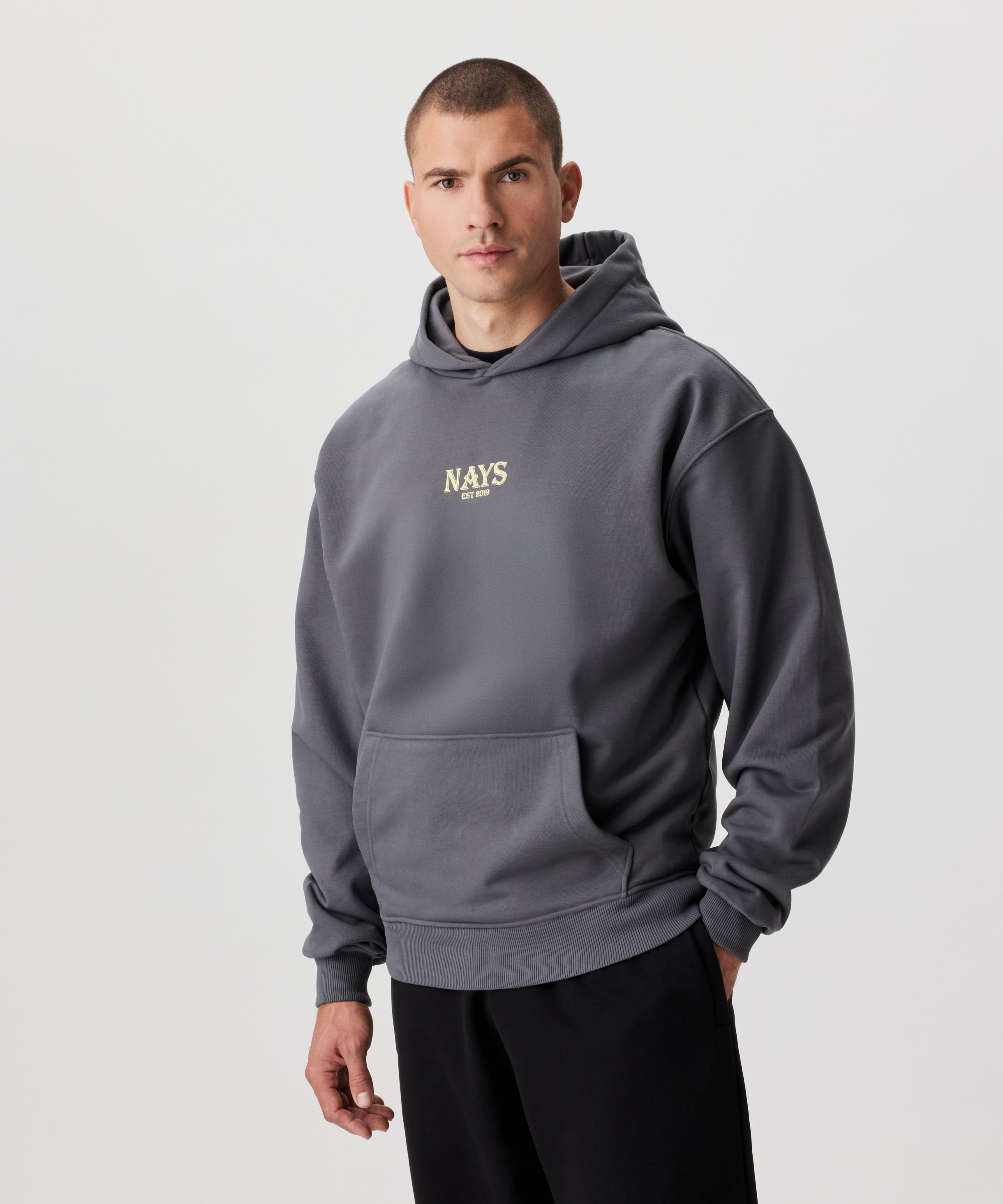 Mann im grauen Kapuzen Sweatshirt in der frontalen Ansicht. Das Sweatshirt hat einen mittig gesetztes Nays-Logo in gelb.