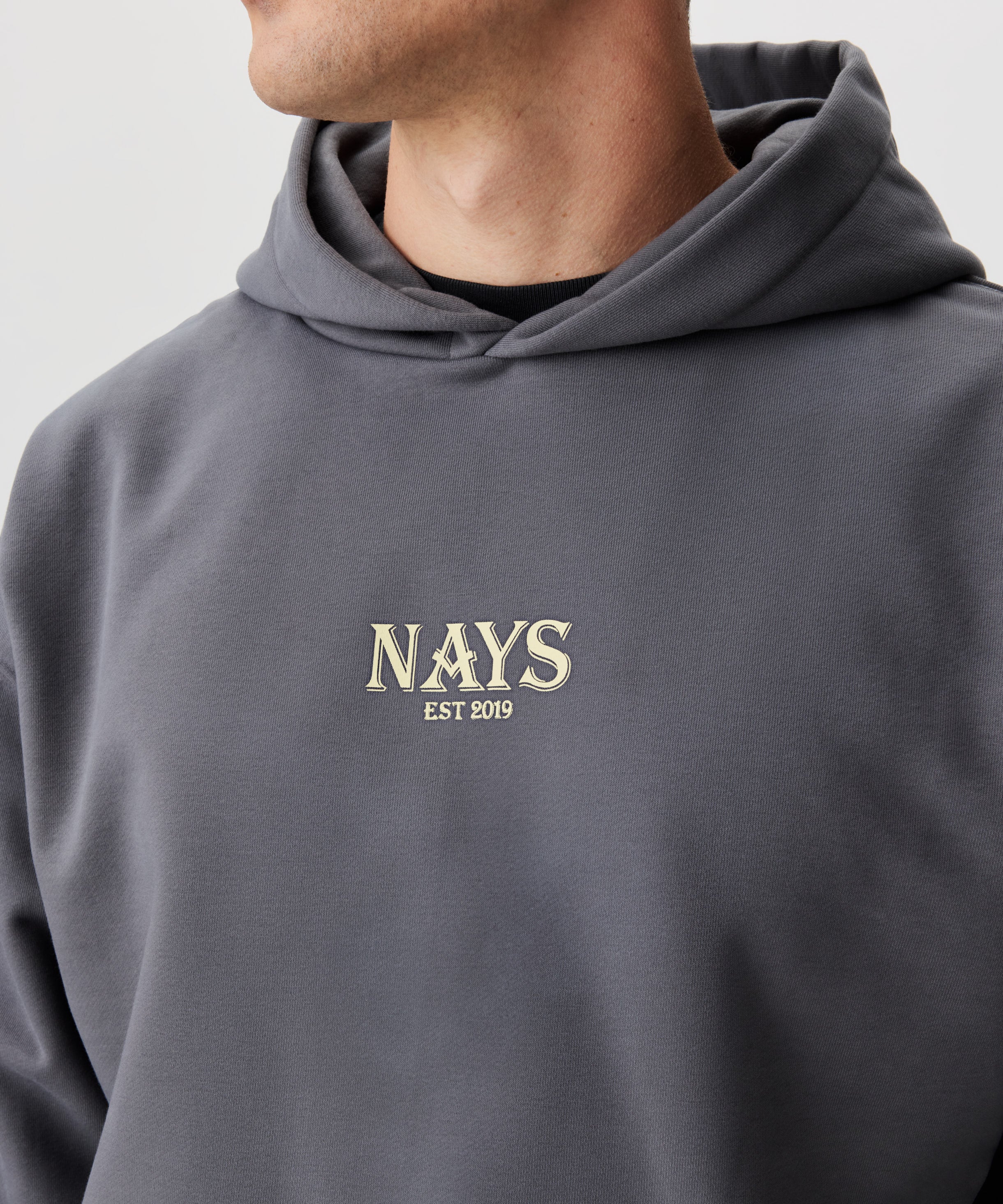Frontansicht Mann im grauen Kapuzen Sweatshirt. Das Sweatshirt hat einen mittig gesetztes Nays-Logo in gelb.