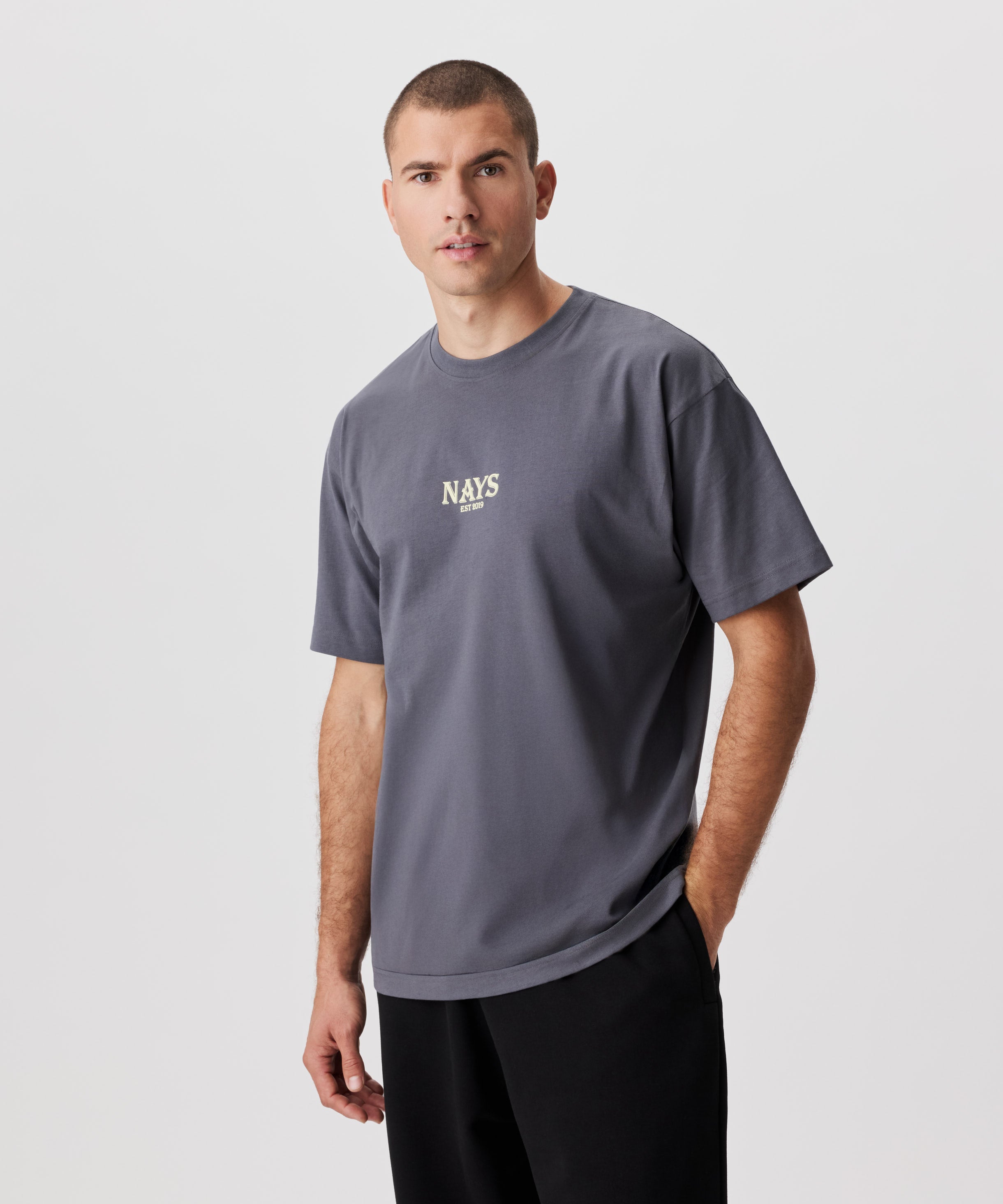 Frontale Ansicht Mann im grauen T-Shirt. Das T-Shirt hat einen mittig gesetztes Nays-Logo in gelb.