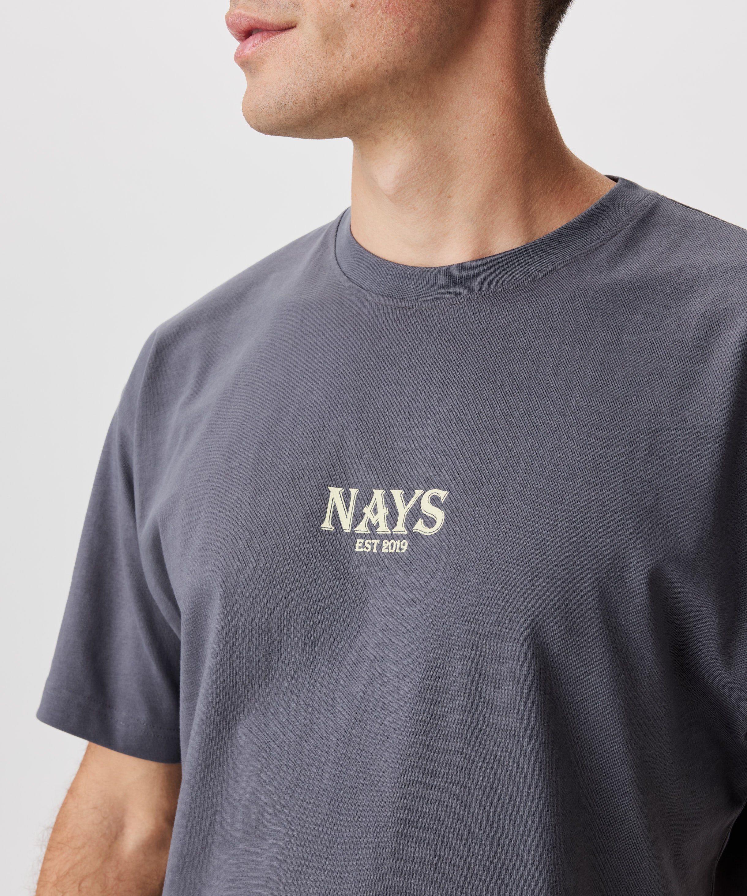 Frontansicht Mann im grauen T-Shirt. Das T-Shirt hat einen mittig gesetztes Nays-Logo in gelb.