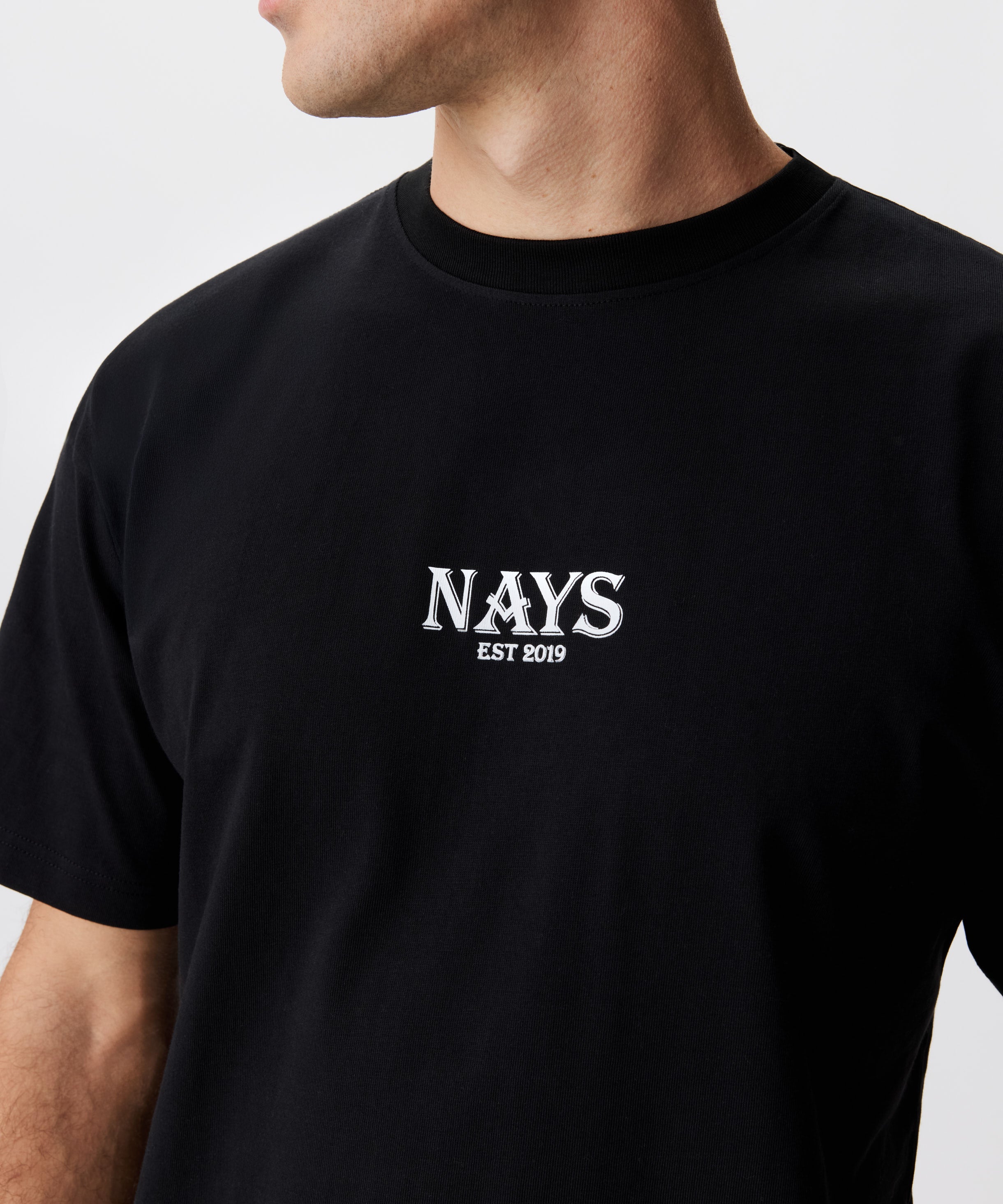 Mann im schwarzen T-Shirt in der frontalen Ansicht. Das T-Shirt hat einen mittig gesetztes Nays-Logo in weiß.
