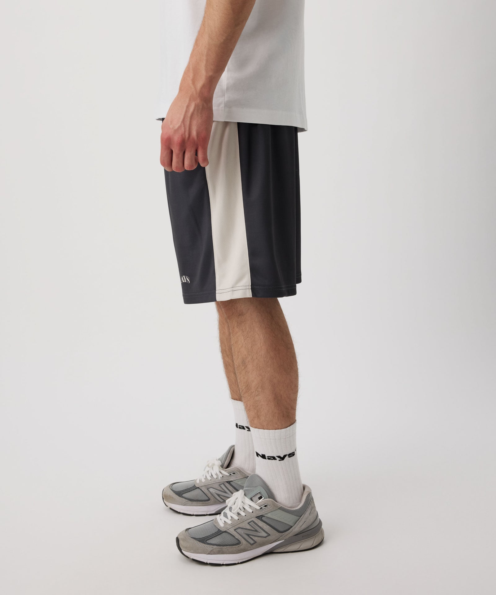 Das Model trägt eine blau-graue-kurze Hose. In der seitlichen Perspektive erkennt man weiße Applikationen an den Seiten. Dazu trägt er ein weißes T-Shirt, helle Socken und graue Turnschuhe.