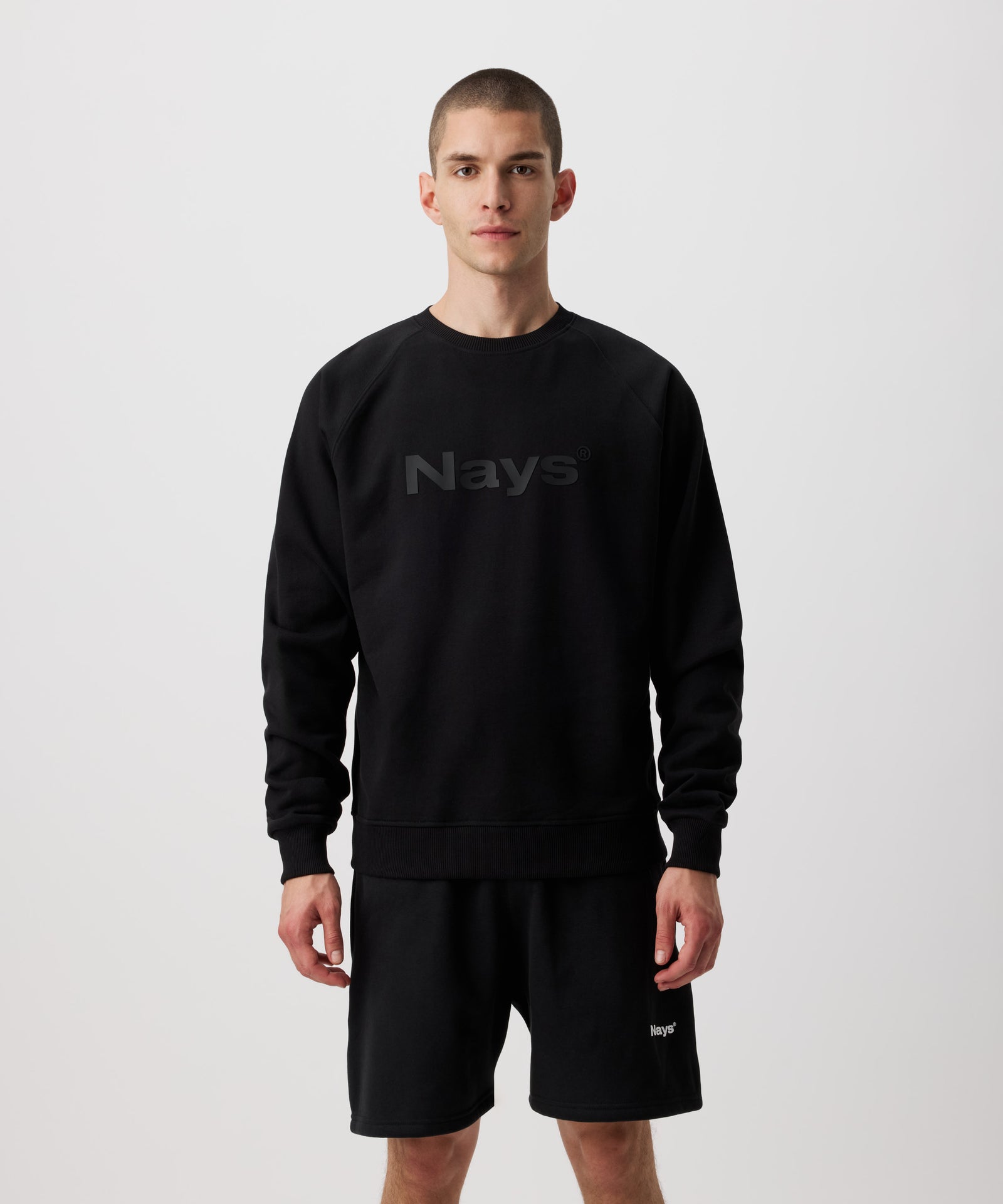 Ein Mann trägt einen schwarzen Pullover. Auf der Brust ein großes, tonales Nays Logo zu sehen. Dazu trägt er eine schwarze Shorts mit weißem Nays Logo Aufdruck.