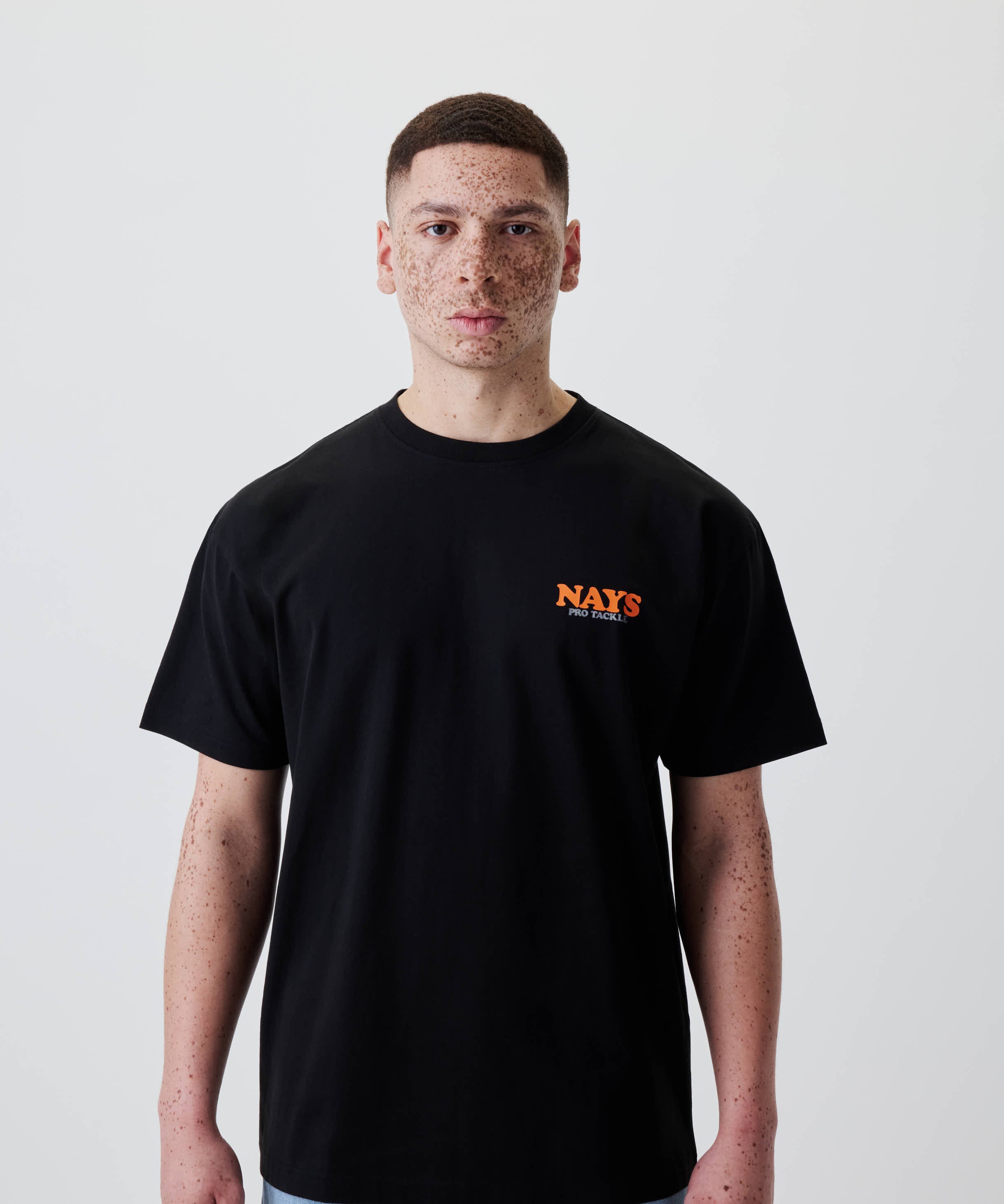 Person mit kurzen Haaren trägt das Nays College Print Tee mit orangefarbenem NAYS Pro Tackle Logo auf der Brust, schlicht und modern gestaltet.