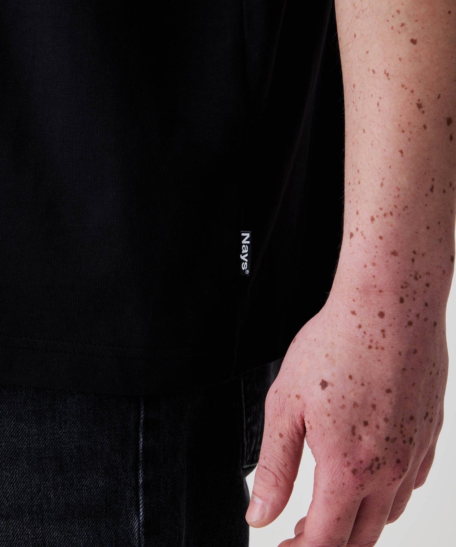 Person trägt das Nays Loose Fit Chest Print Tee mit kleinem Flag Label am Saum, kombiniert mit dunklen Jeans. Frecklige Hand sichtbar gegen hellen Hintergrund.