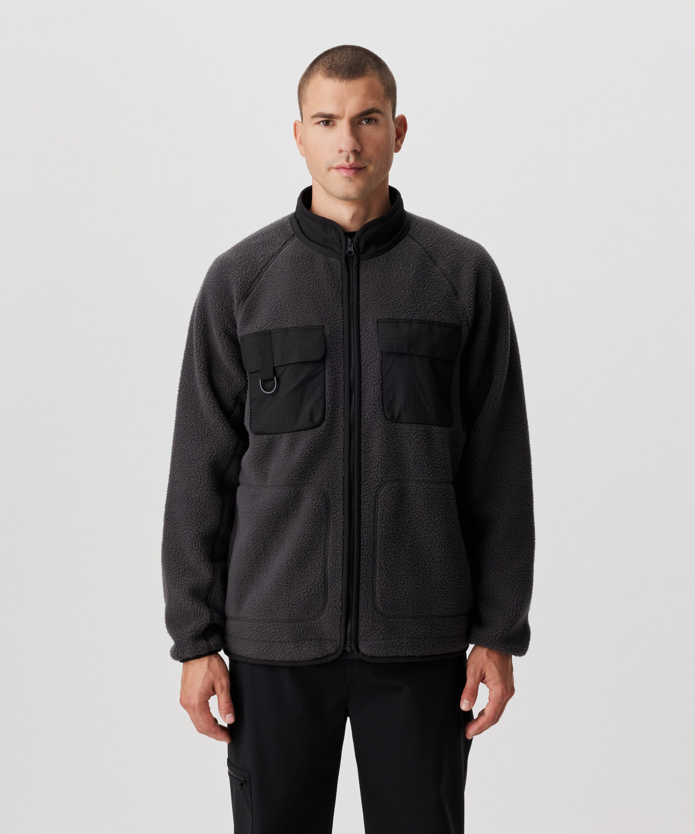 Frontansicht von einem Mann in dunkel-grauer Fleece-Jacke mit abgesetzten Kragen und Brusttaschen im schwarzen Ripstop. Die Jacke verfügt über zwei Brust- und zwei weitere, seitliche Taschen.