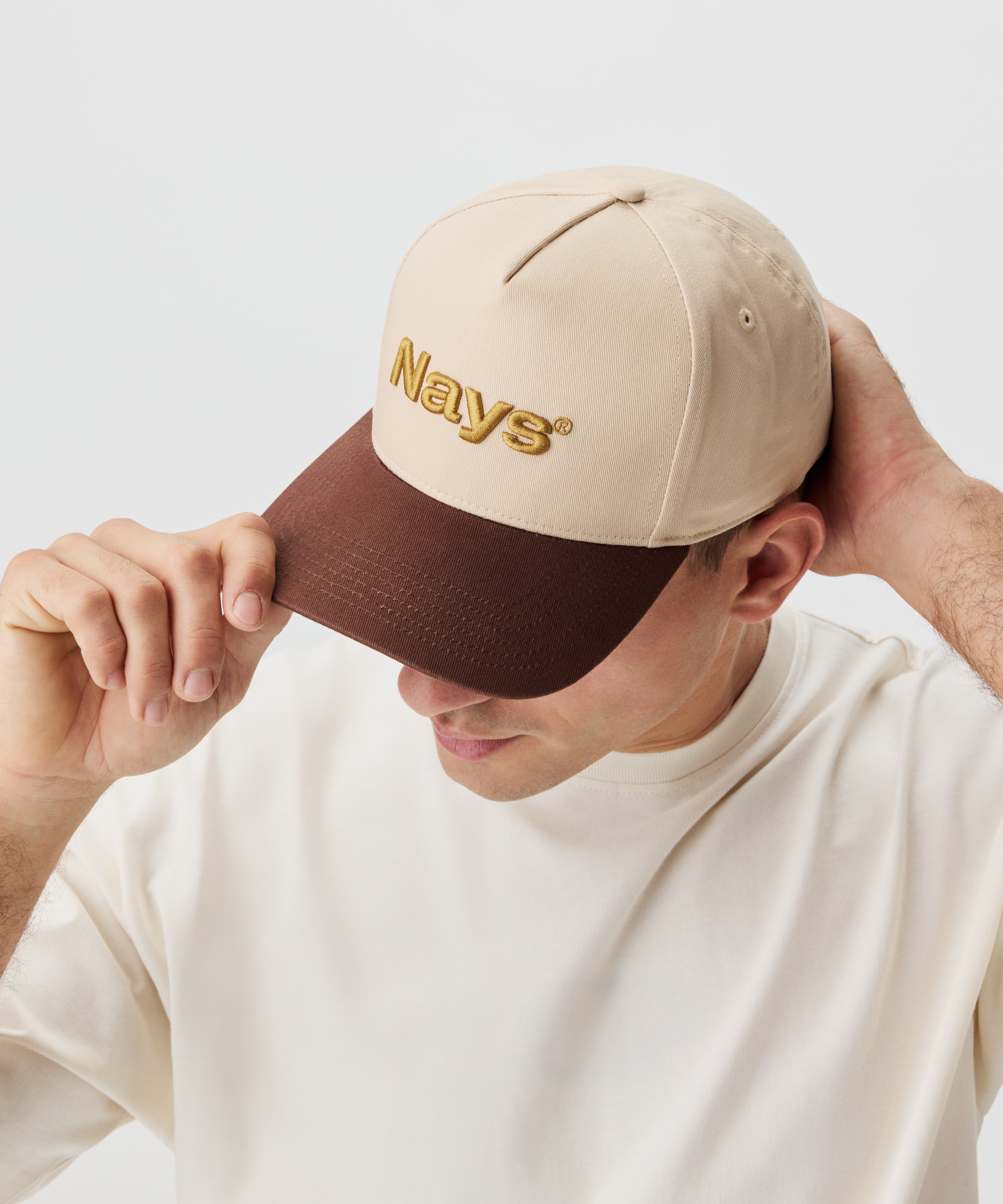 Ein Mann setzt eine Cap auf. Die Cap ist beige mit einem braunen Schirm und gold-gelbem Nays-Logo Strick.