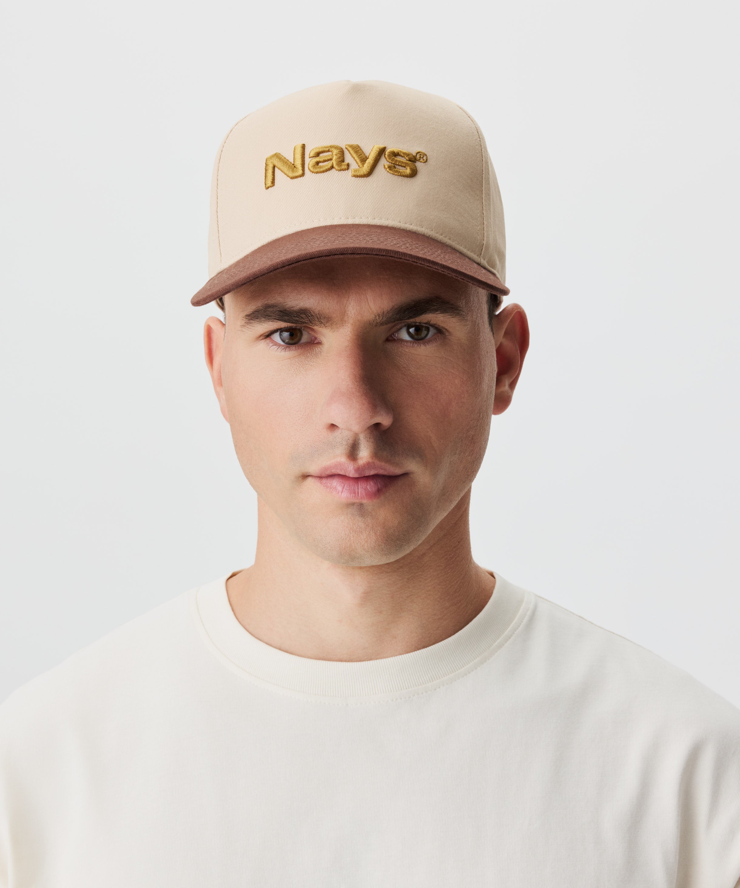 Frontansicht Mann mit Cap. Die Cap ist beige mit einem braunen Schirm und gold-gelbem Nays-Logo Strick.