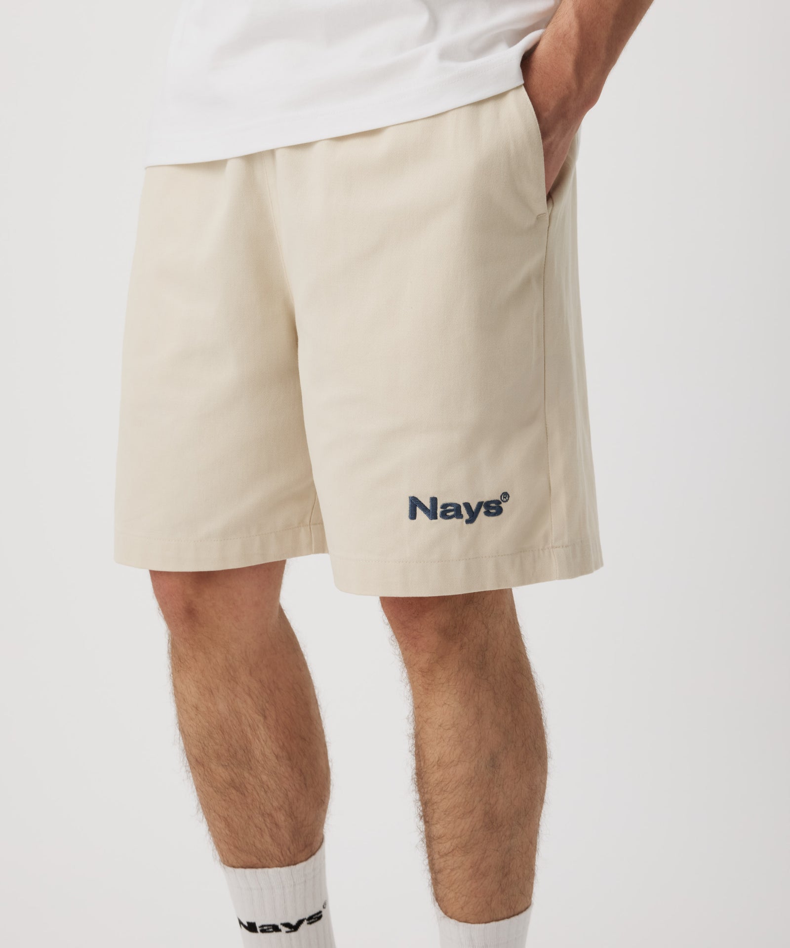 Ein Mann trägt eine beige Shorts mit einem dunkel-grünen "Nays"-Logo auf dem linken Bein und hat seine linke Hand in der Tasche. Dazu trägt er ein weißes T-Shirt und eine weiße Socken.