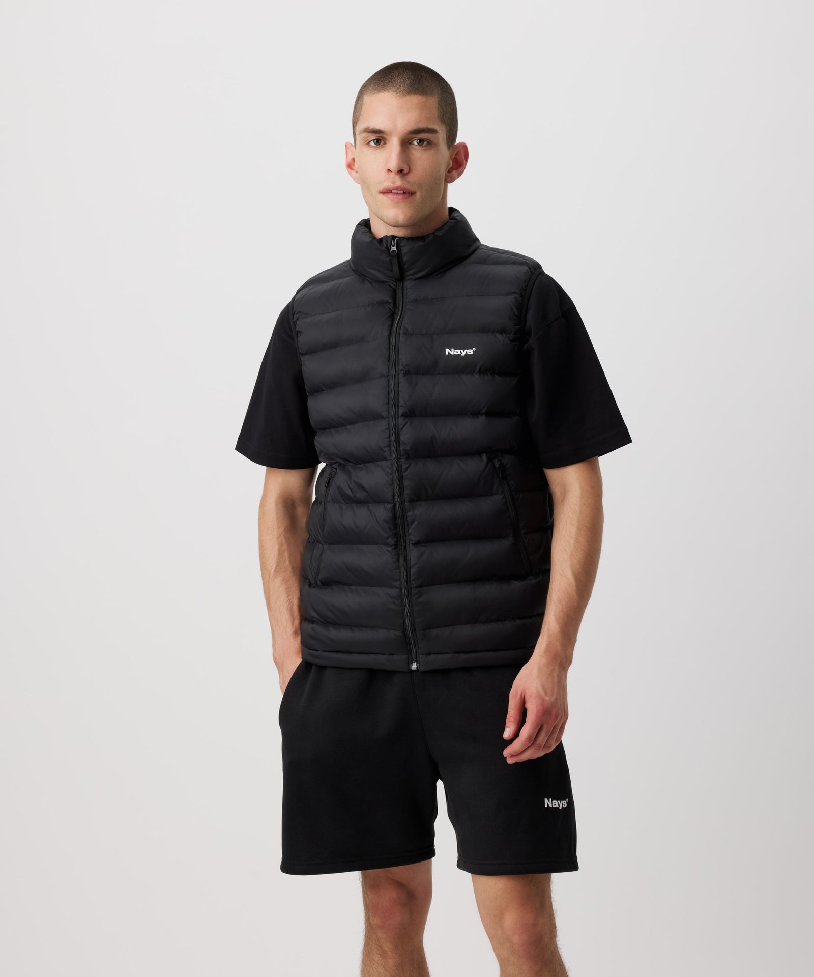 Ein Mann trägt eine schwarze Steppweste, eine schwarze Shorts und ein schwarzes T-Shirt.