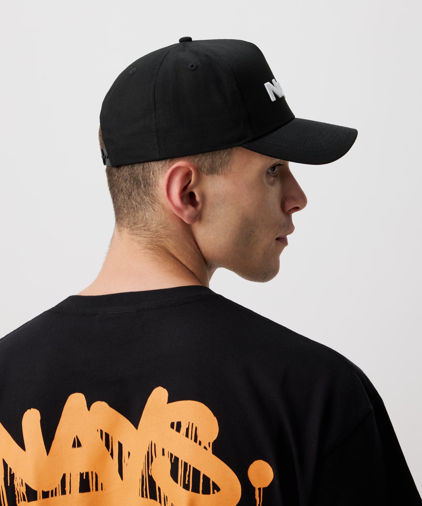 Ein Mann im Seitenprofil trägt eine schwarze Schirm Mütze. Dazu trägt er ein schwarzes T-Shirt mit orange-farbigem Nays Logo.