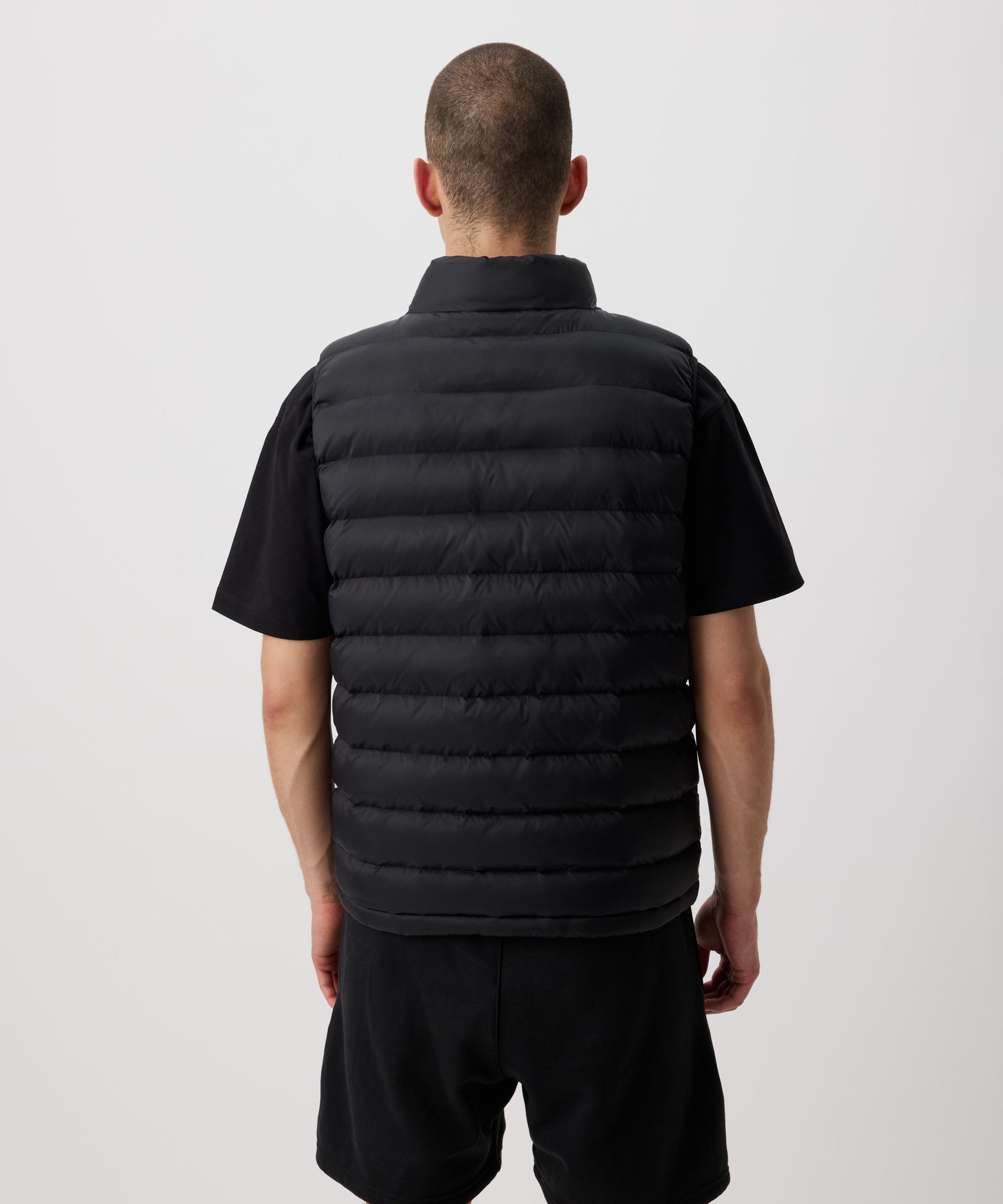 Ein Mann von hinten zu sehen - er trägt eine leichte Steppweste in schwarz und eine kurze schwarze Hose.