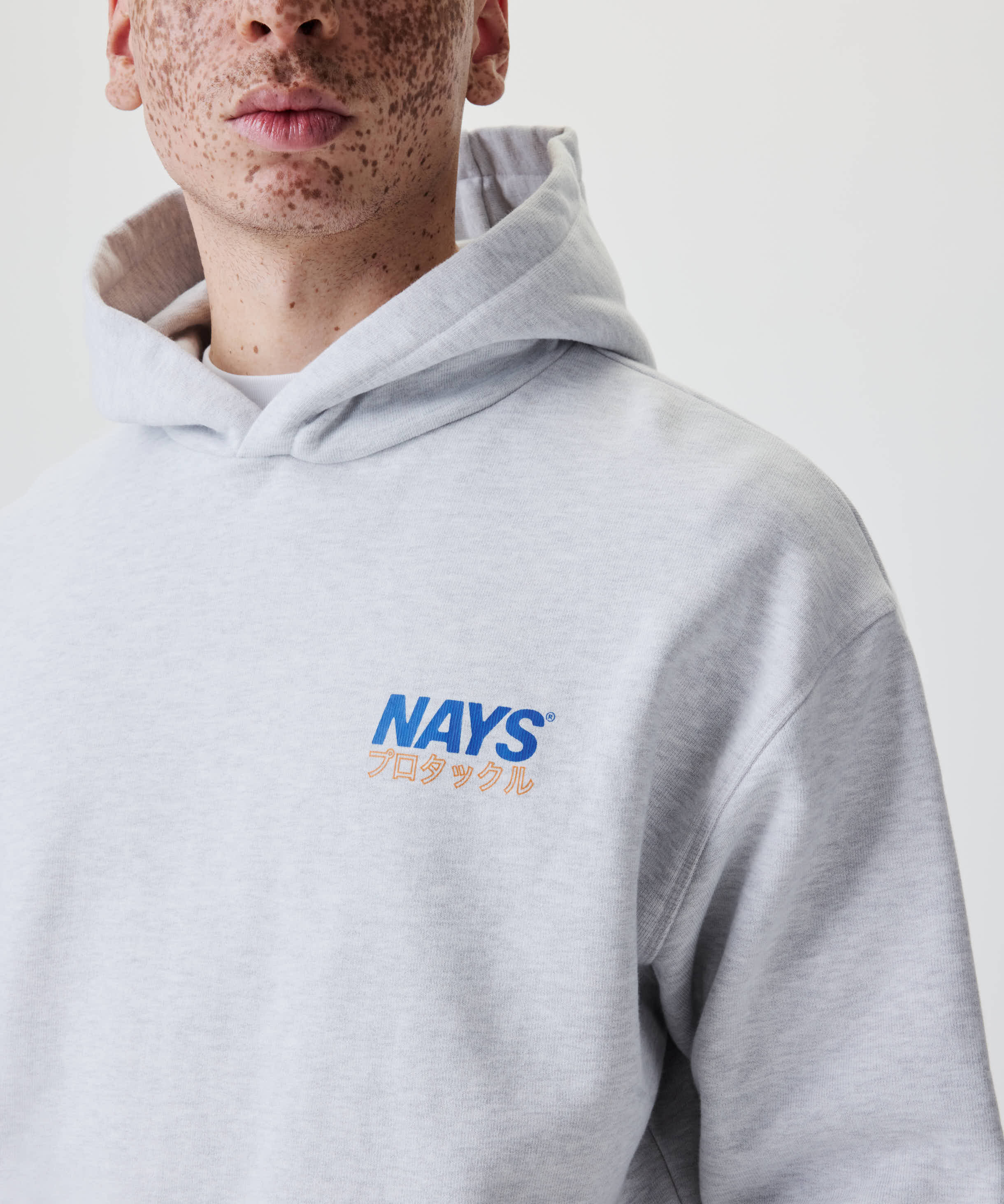 Person trägt das Nays Pro Tackle International Hoodie mit kleinem blauen Logo und orangen Katakana-Zeichen auf der Brust, Kapuze um die Schultern.