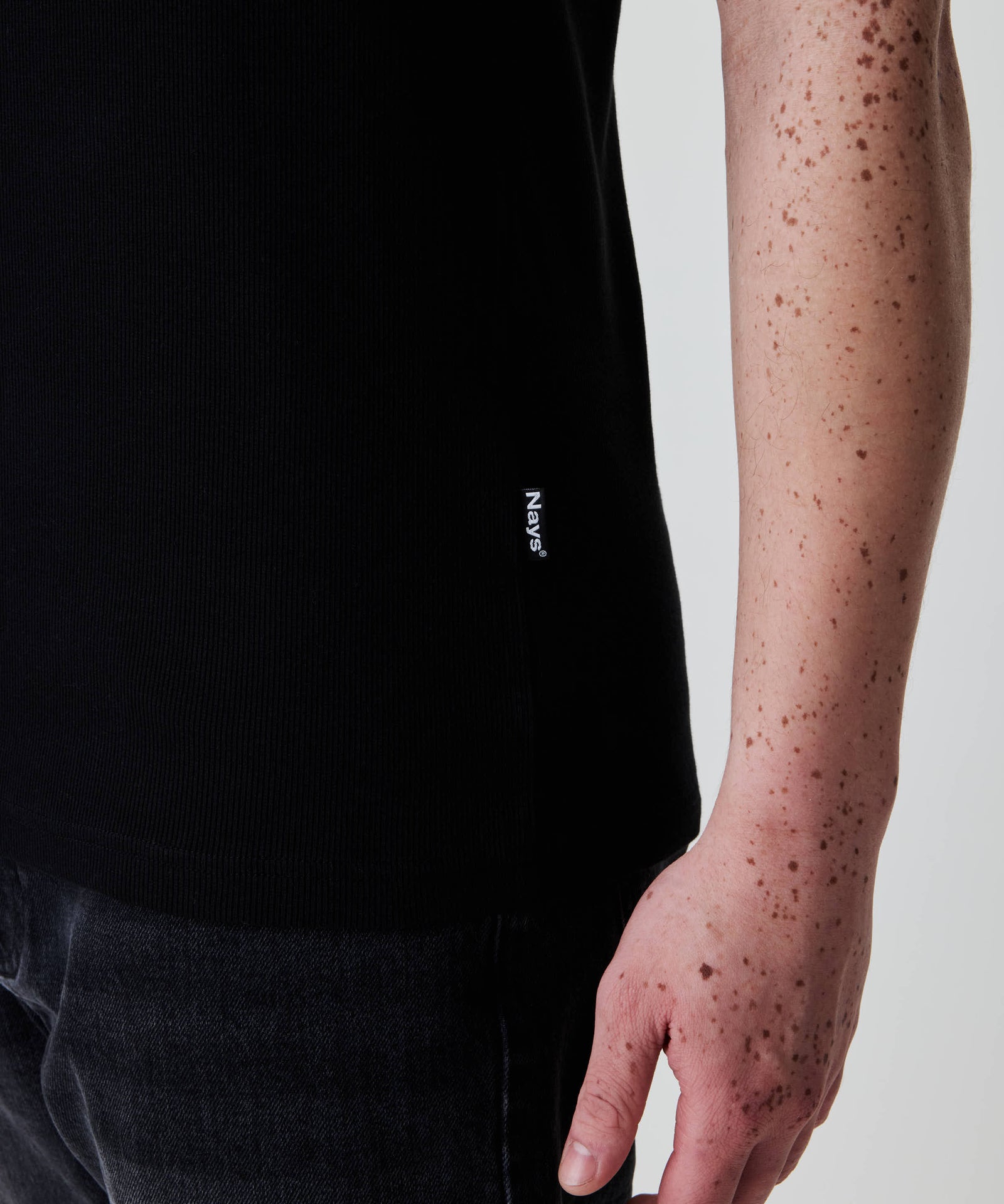 Person trägt das Nays Classic Rib Tank Top in Schwarz mit kleinem Label am Saum, kombiniert mit dunklen Jeans. 