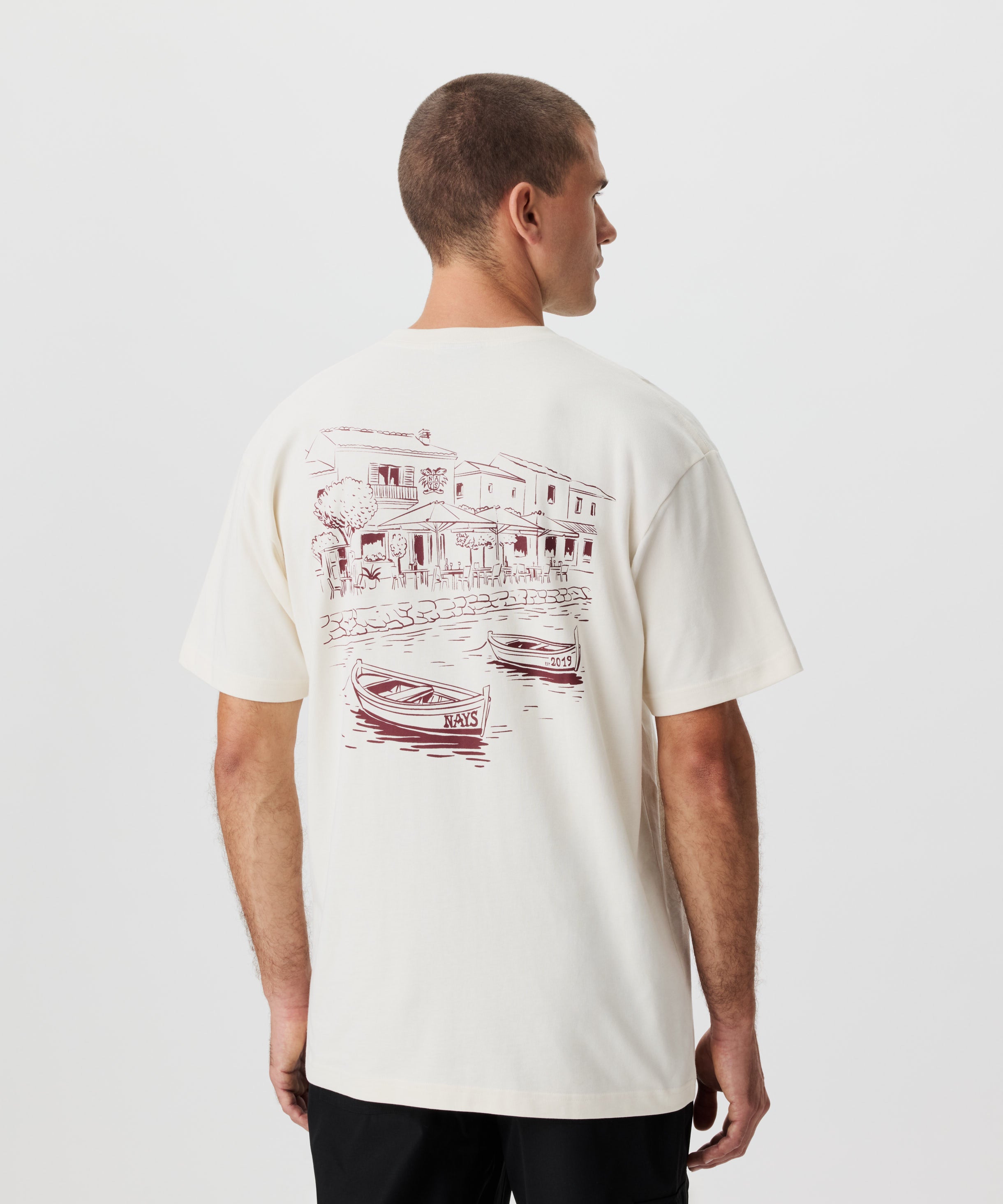 Rückansicht Mann im beigen T-shirt und schwarzer Hose. Das T-Shirt weist einen großen Rückenprint in dunkelrot auf.