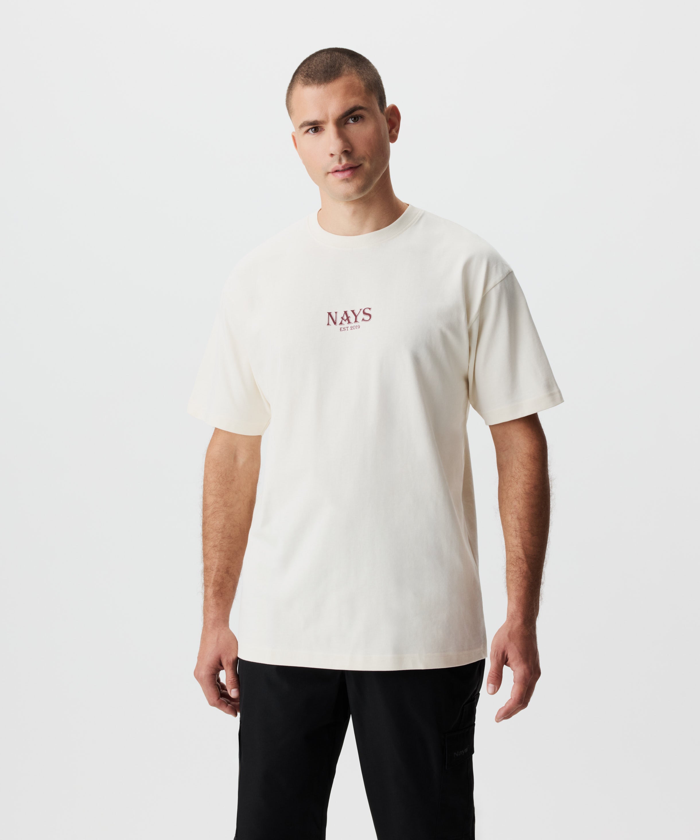 Frontansicht Mann im beige-farbigem T-sShirt und schwarzer Hose. Das T-Shirt hat einen Nays-Logo-Schriftzug auf der Brust.