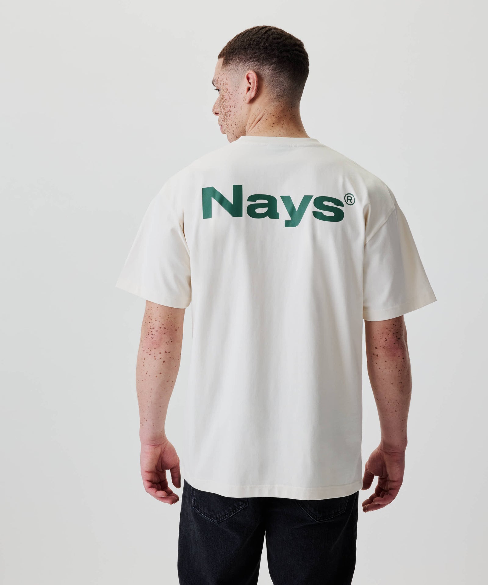 Person von hinten mit Nays Quote Tee im lockeren Schnitt, dunkelgrüne Grafik auf dem Rücken, freiliegende Arme mit Sommersprossen, kombiniert mit dunklen Hosen.