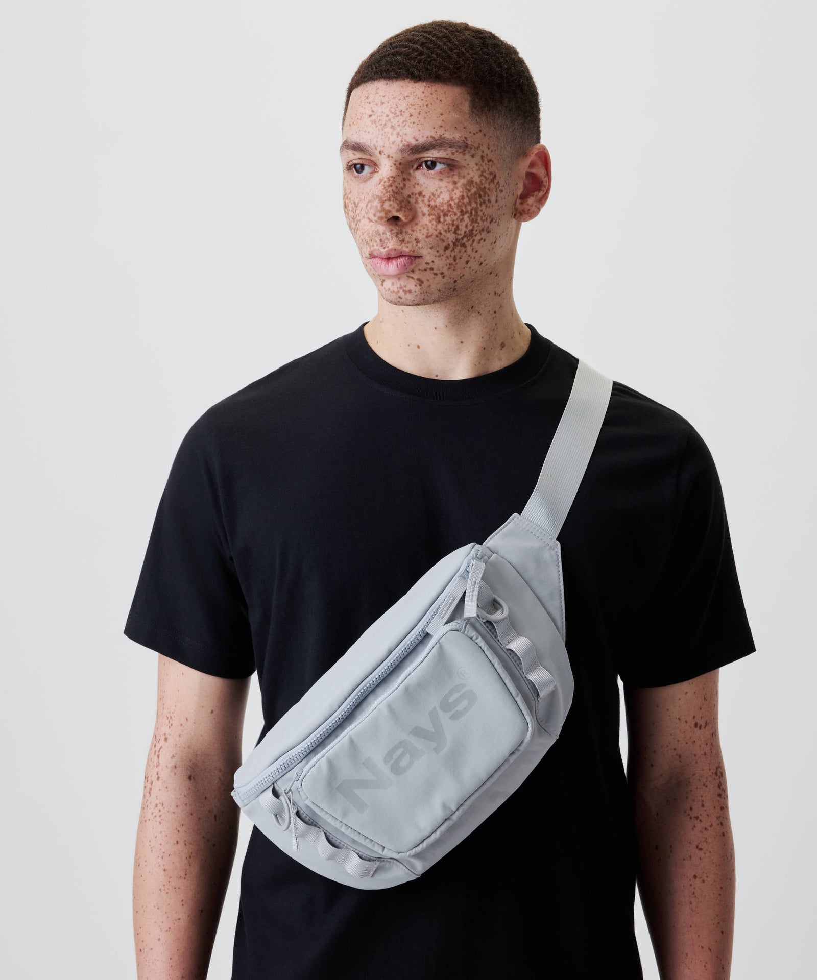 Ein junger Mann trägt die Nays Lightweight Cross Body Bag mit zwei Fächern, verstellbarem Gurt und Ton-in-Ton Logo quer über der Brust.