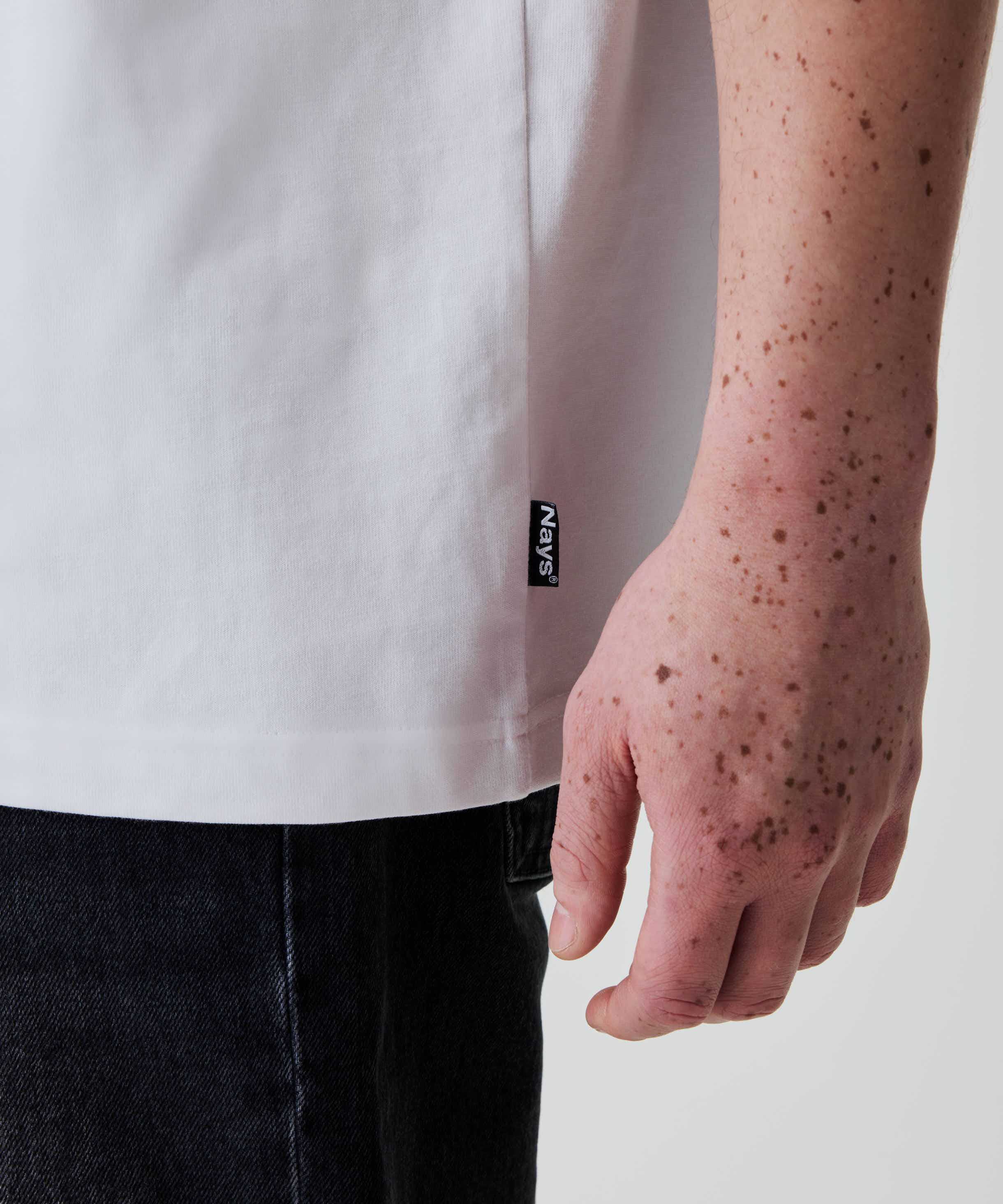 Nays Loose Fit Chest Print Tee in Weiß, locker sitzend, mit sichtbarem Nays®-Seitenetikett an der Hüfte. Freckles auf Unterarm und Hand des Models.