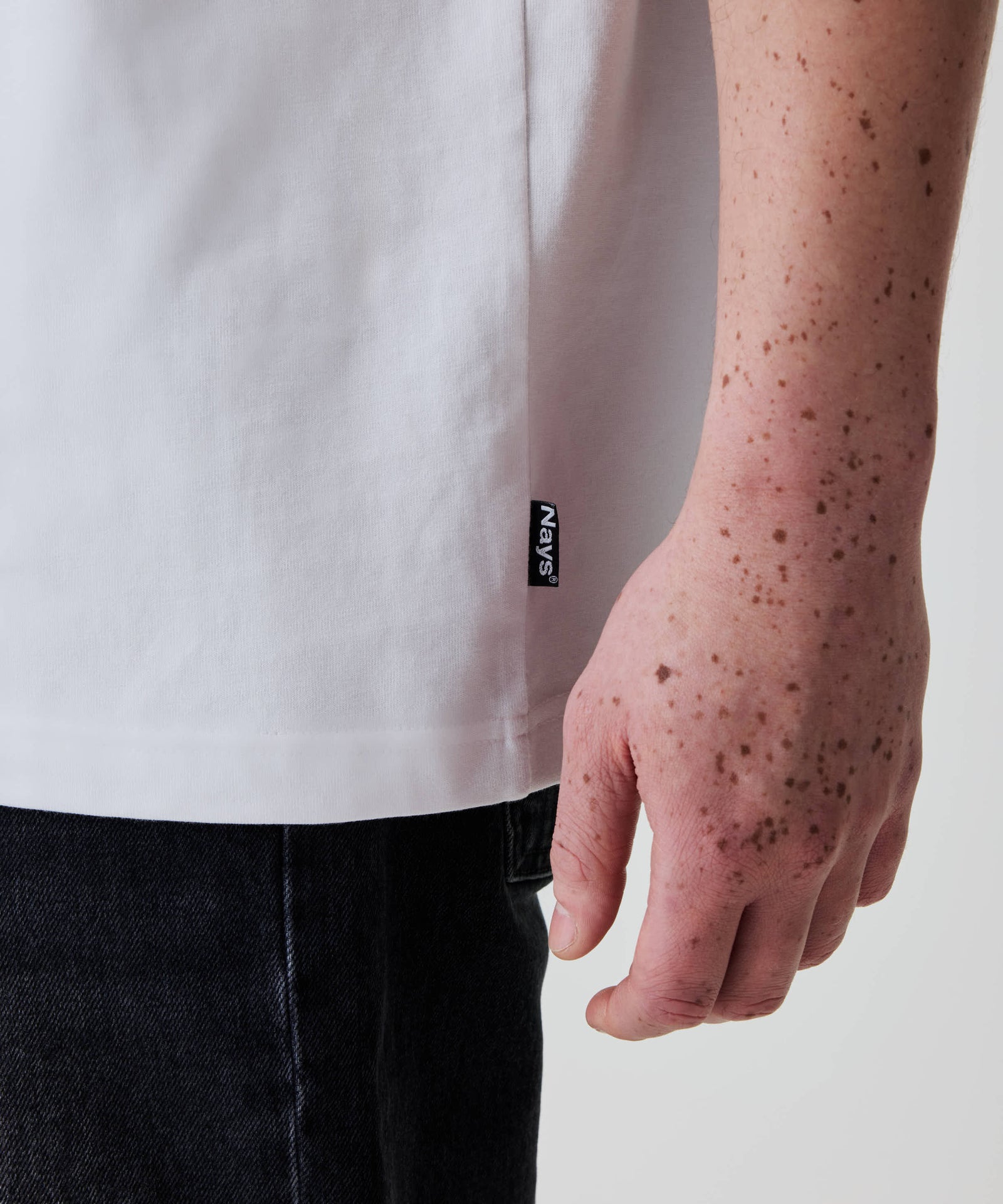 Nays Loose Fit Chest Print Tee in Weiß, locker sitzend, mit sichtbarem Nays®-Seitenetikett an der Hüfte. Freckles auf Unterarm und Hand des Models.